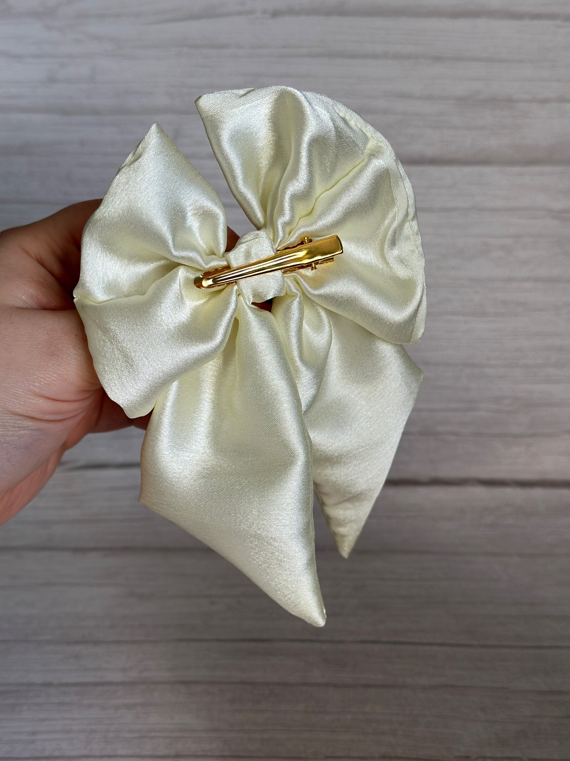 In a hand, there's a shimmering Silk Sailor Bow Clip - Ivory from Crafts by Kate, featuring a gold crocodile clip on the back. The background is a light wooden surface.