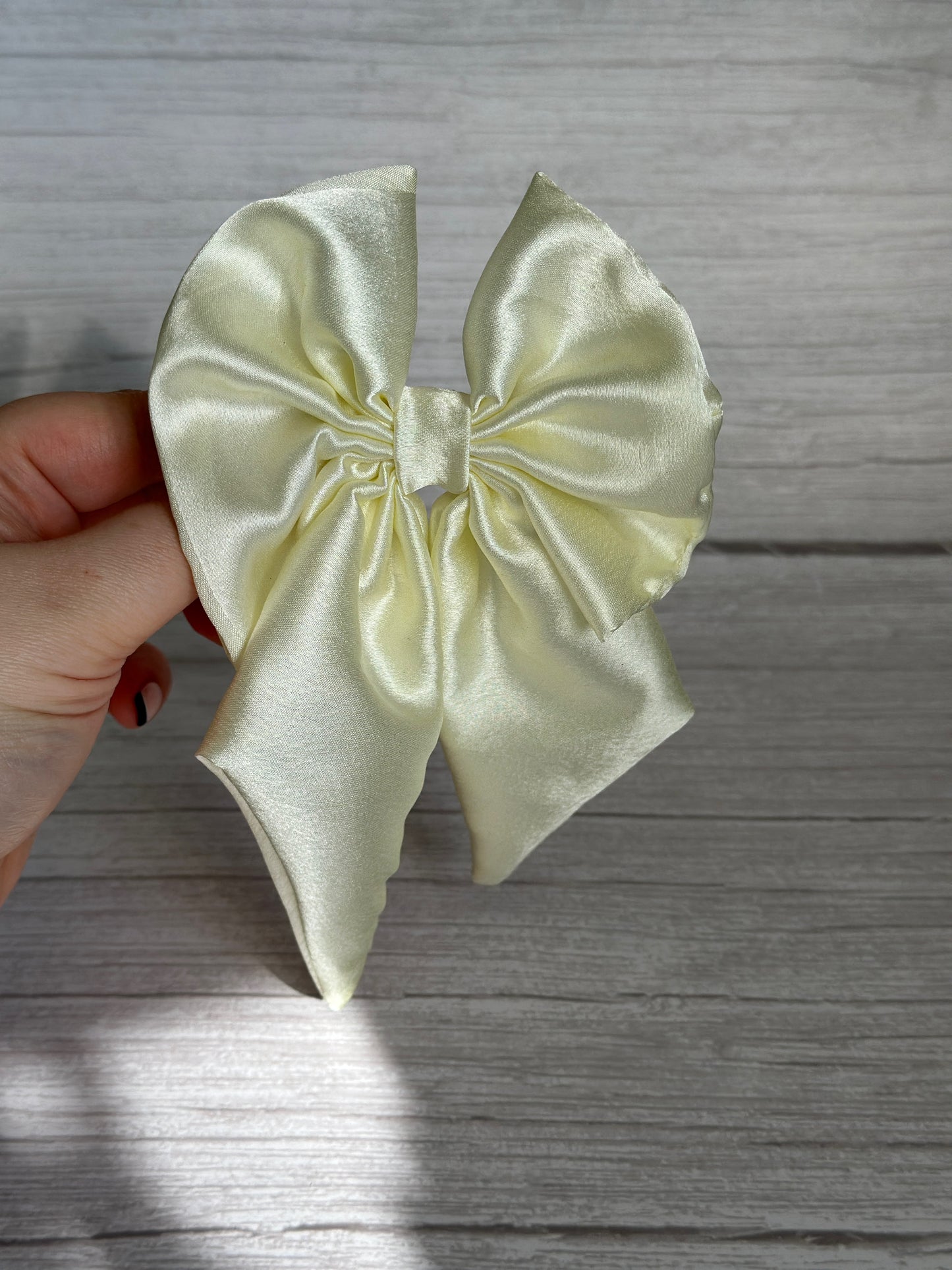 A hand holds an ivory Silk Sailor Bow Clip by Crafts by Kate against a light wooden background. Neatly tied and glossy from the satin material, this handcrafted luxury piece is perfect for adding elegance to any outfit.