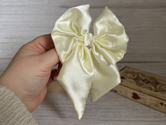 A hand holding the Silk Sailor Bow Clip - Ivory from Crafts by Kate against a wooden background, accompanied by a decorative box adorned with heart patterns, with the clip gleaming in soft cream tones.