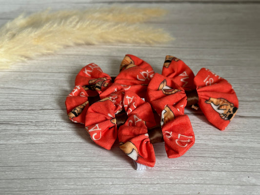 Several small, handcrafted Cotton Pet Bows with collar attachments, from the "Karma is a Cat" collection by Crafts by Kate, featuring a red background adorned with dog illustrations are arranged on a light wooden surface. There's a fuzzy light-colored decoration in the top left corner of the image, adding to the charm of these adorable accessories.