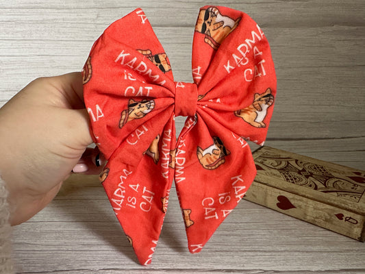 A hand holds up the Crafts by Kate Cotton Sailor Bow Clip, a large red fabric bow adorned with illustrations of cats wearing scarves and the text "KARMA IS A CAT." The bow is showcased in front of a wooden surface and a decorative wooden box featuring spade and heart designs.