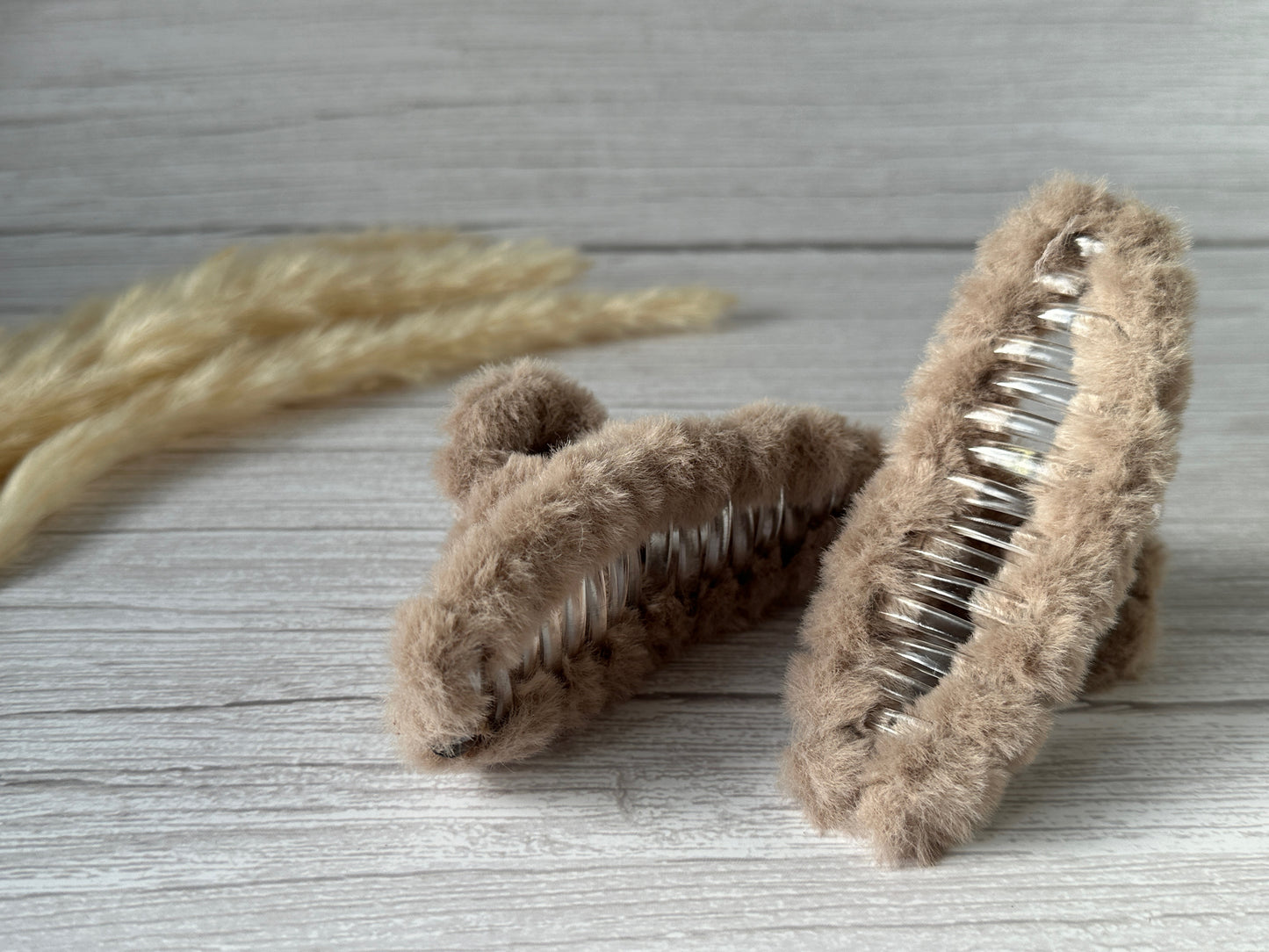 Two Crafts by Kate Fluffy Hair Claw Clips in Latte Brown, covered in soft, plush fabric, are elegantly displayed on a light gray wooden surface. Beige dried wheat stalks in the background enhance the natural appeal of the scene.