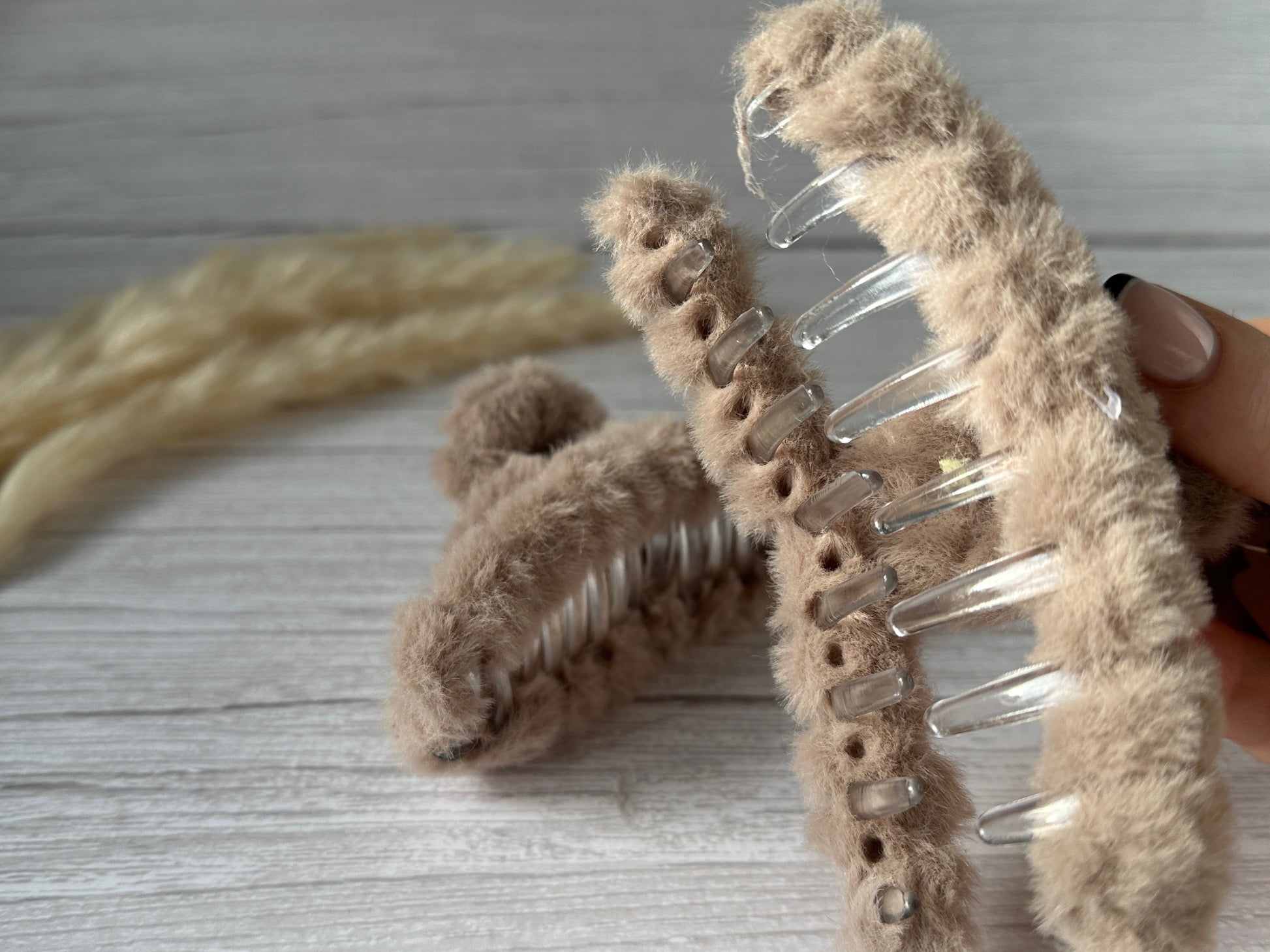 A close-up of a hand holding a large, handcrafted Fluffy Hair Claw Clip - Latte Brown by Crafts by Kate, showcasing its transparent comb-like teeth. Another luxury plush fabric hair clip lies on a wooden surface in the background. Both clips have a soft, fluffy texture and are light brown in color.