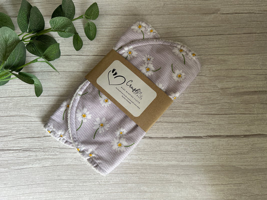 A set of Crafts by Kate's Cotton & Terry Towelling Baby Burping Cloths, featuring a lavender daisy motif on a light purple background, encased in cardboard packaging labeled with "CareCloth." They are displayed on a wooden surface next to green leaves, embodying both elegance and eco-friendliness.