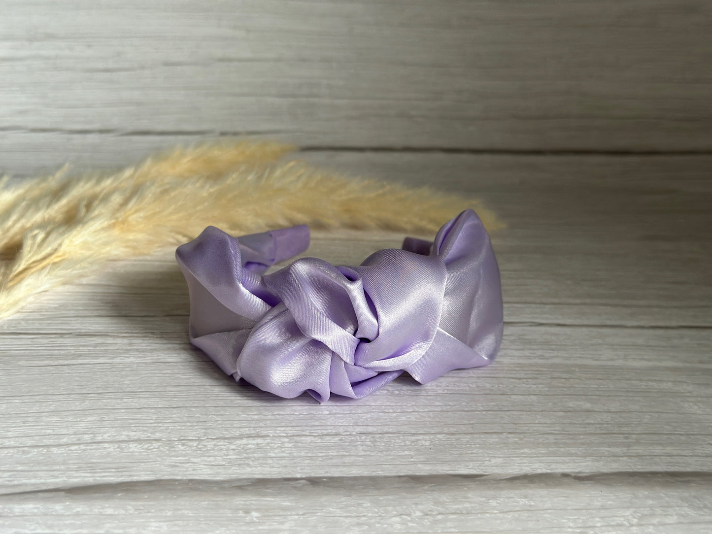 A Silk Knotted Headband in Lavender Purple from Crafts by Kate graces a light wooden surface, exuding elegance. In the backdrop, delicate beige pampas grass enhances its charm.