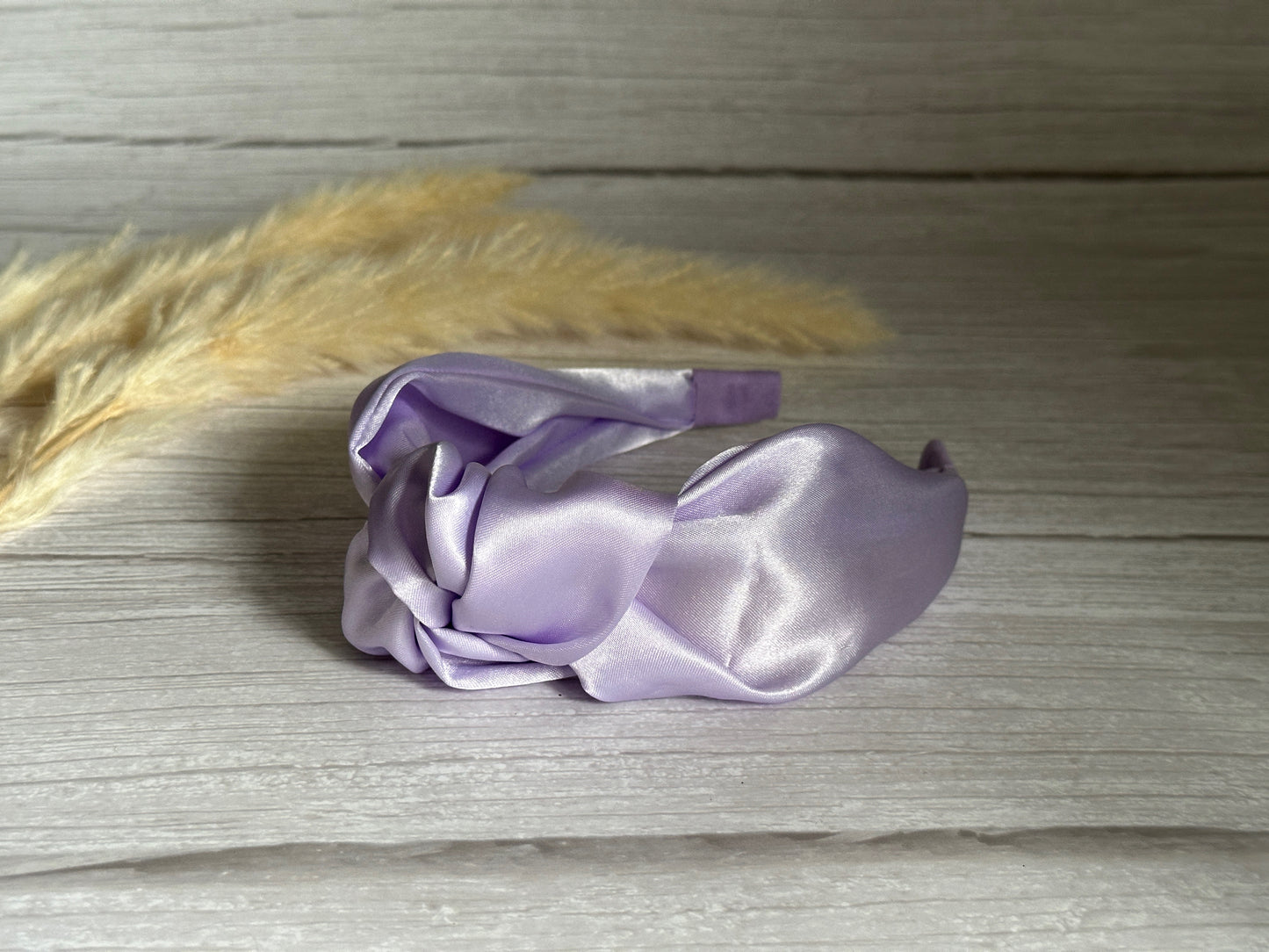 The Crafts by Kate Silk Knotted Headband in Lavender Purple rests elegantly on a light wooden surface, with blurred pampas grass softly framing the background.