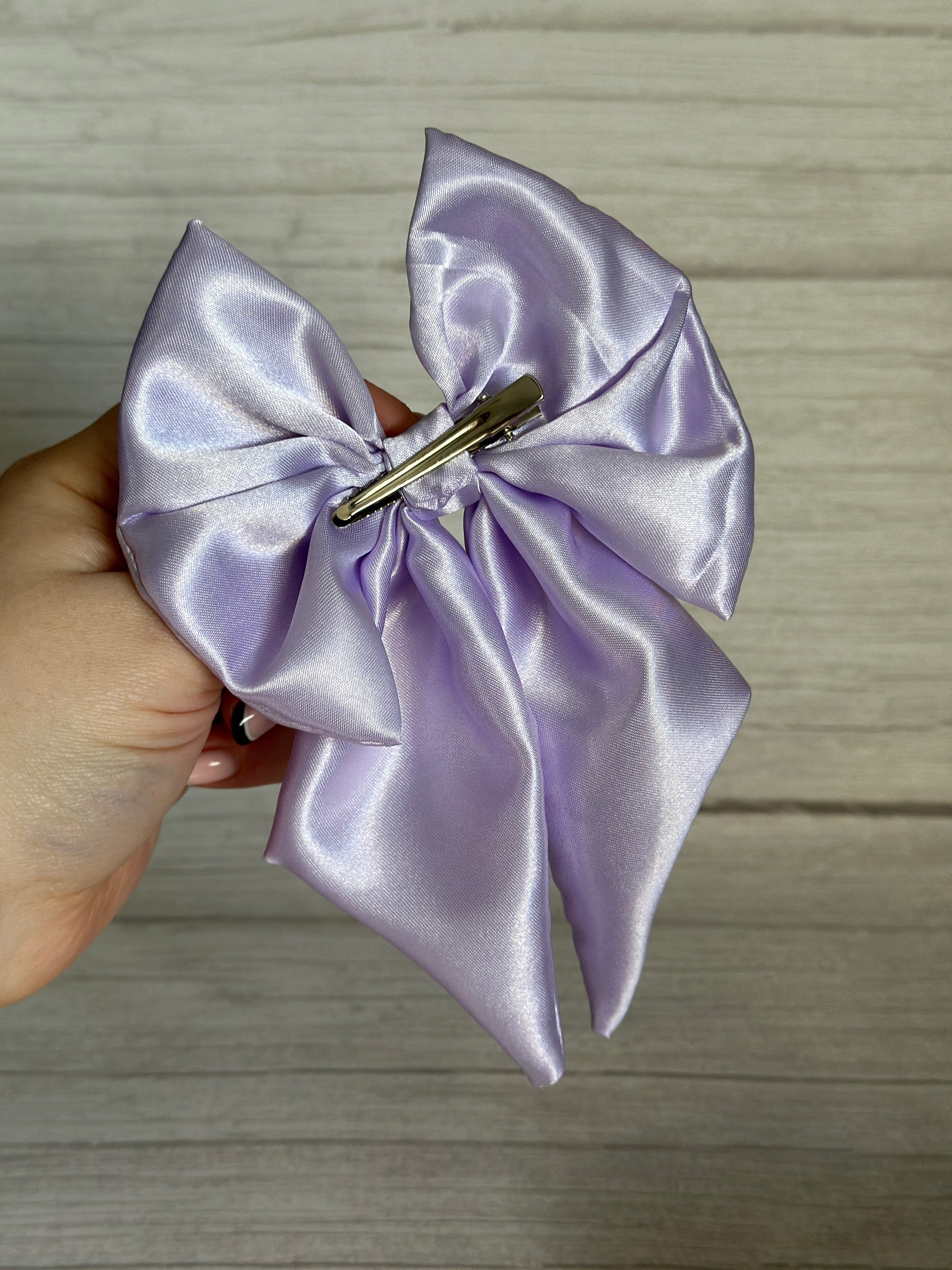 A hand holds the exquisite Silk Sailor Bow Clip in Lavender Purple from Crafts by Kate, its lustrous silk fabric secured with a crocodile clip, set against a light wooden background.