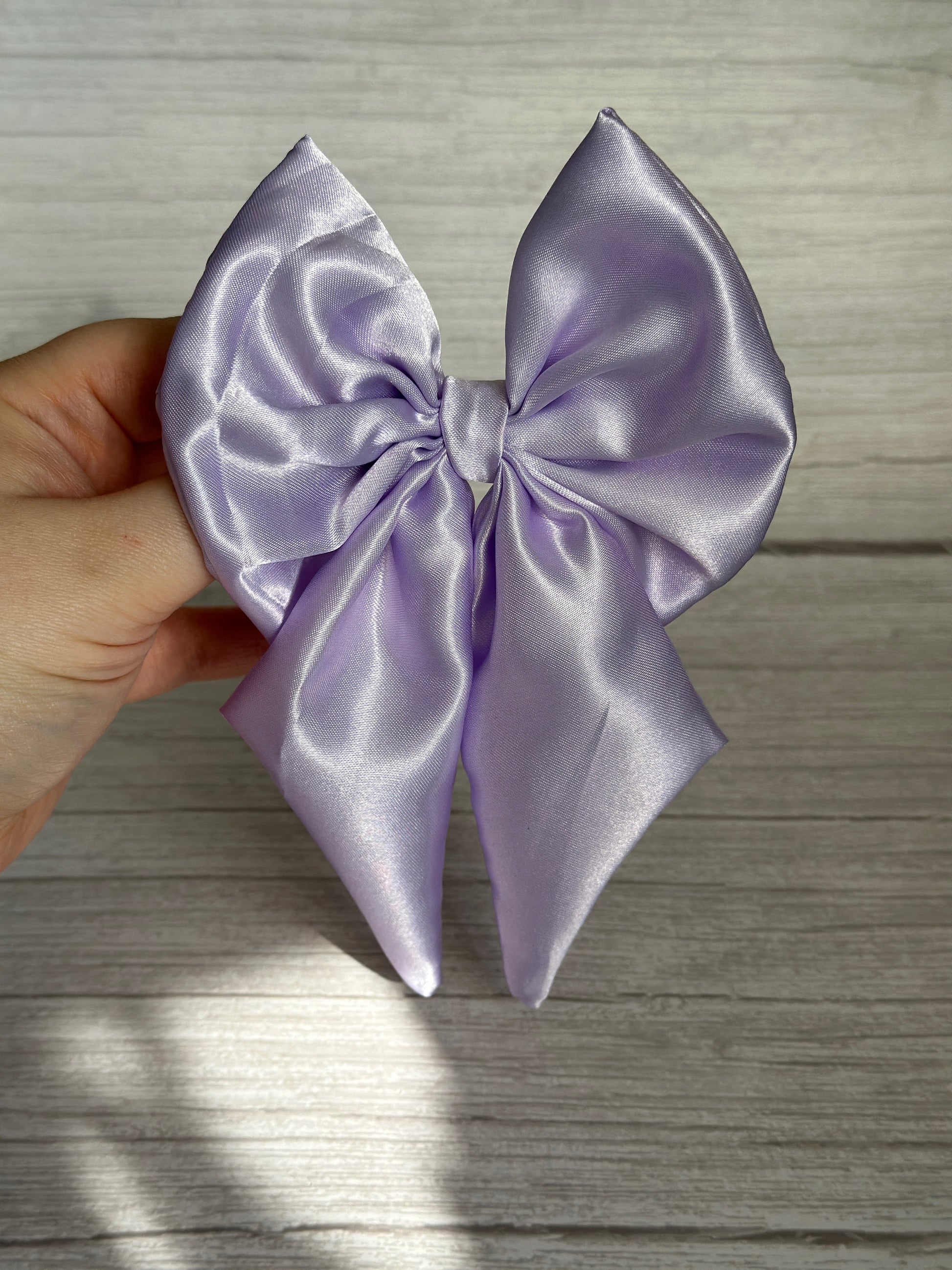 A hand holds the Crafts by Kate Silk Sailor Bow Clip in shimmering lavender purple against a light wooden background. Its smooth, satin-like texture catches the light, highlighting elegant folds, and it's secured with a sleek crocodile clip.