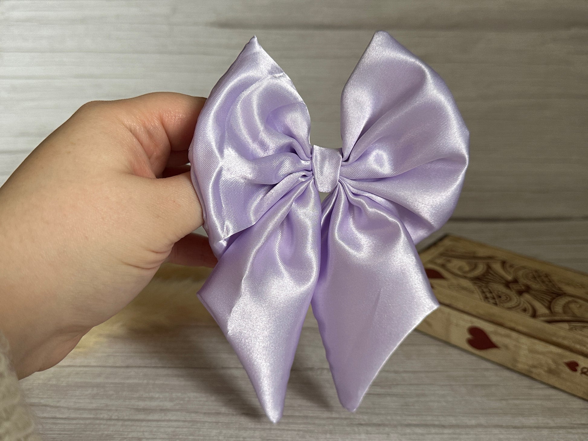 A hand holds the luxurious Silk Sailor Bow Clip in lavender purple by Crafts by Kate against a wood-textured background. The bow features a symmetrical design with two loops and two tails, made from silk fabric and secured with a discreet crocodile clip.