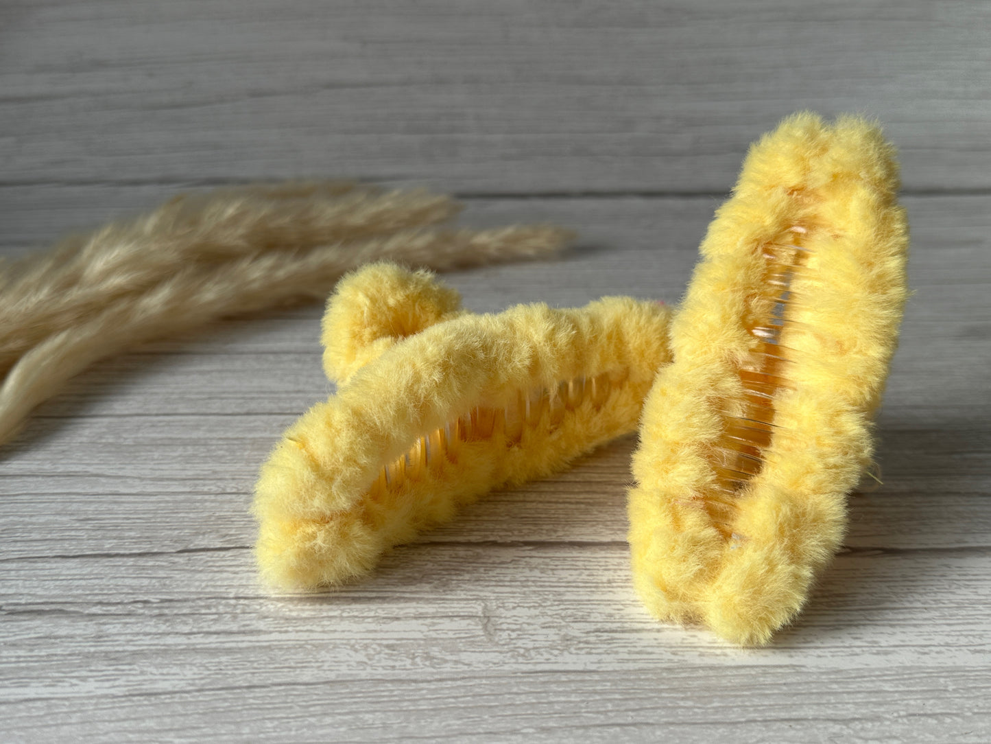 Two Crafts by Kate "Fluffy Hair Claw Clips" in lemon yellow are placed on a light wooden surface. The clips have a soft, luxury plush fabric exterior with visible inner grips. In the background, there are some dried, light brown pampas grass plumes.