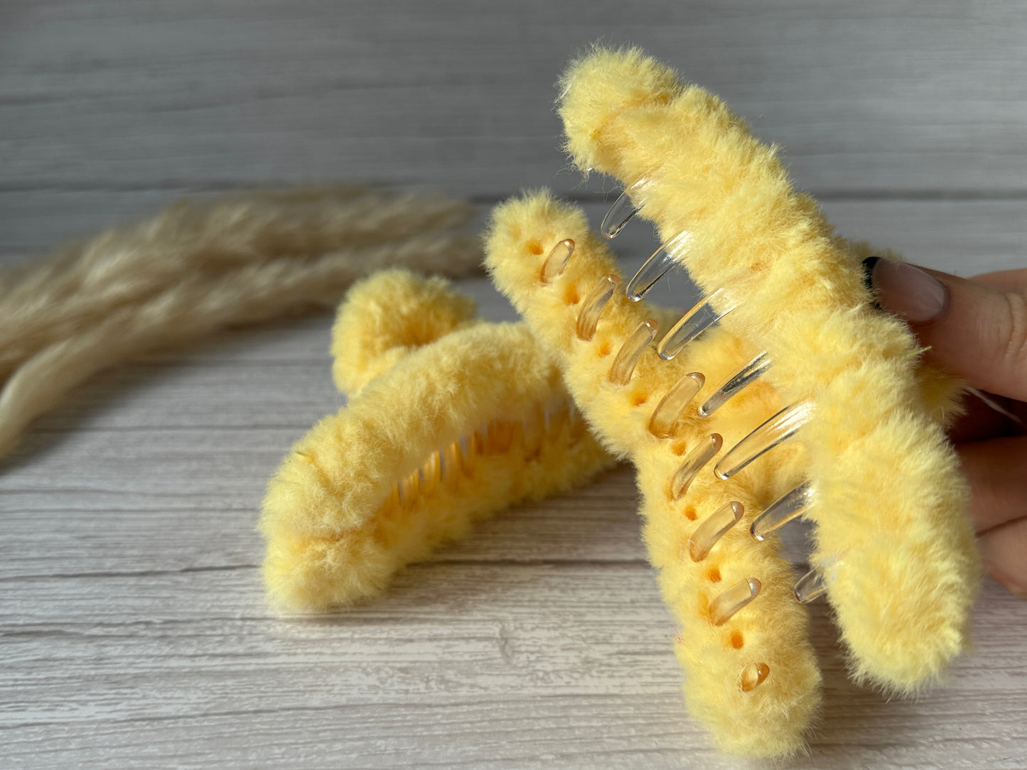 A hand is holding two Crafts by Kate "Fluffy Hair Claw Clips" in Lemon Yellow, crafted from luxury plush fabric, against a light wood background. The clips have a soft, plush texture with metal mechanisms underneath. Some grainy beige plant stalks are blurred out in the background.