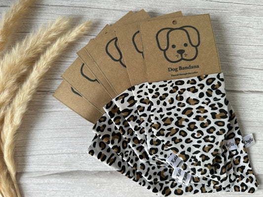 A stack of handcrafted Crafts by Kate Cotton Pet Bandanas with collar attachments and a leopard print design is displayed on a light wooden surface. The bandanas are packaged with a brown cardboard label featuring an illustration of a dog's face and the text "Dog Bandana" printed on it. Dried plants are placed beside this charming pet accessory.