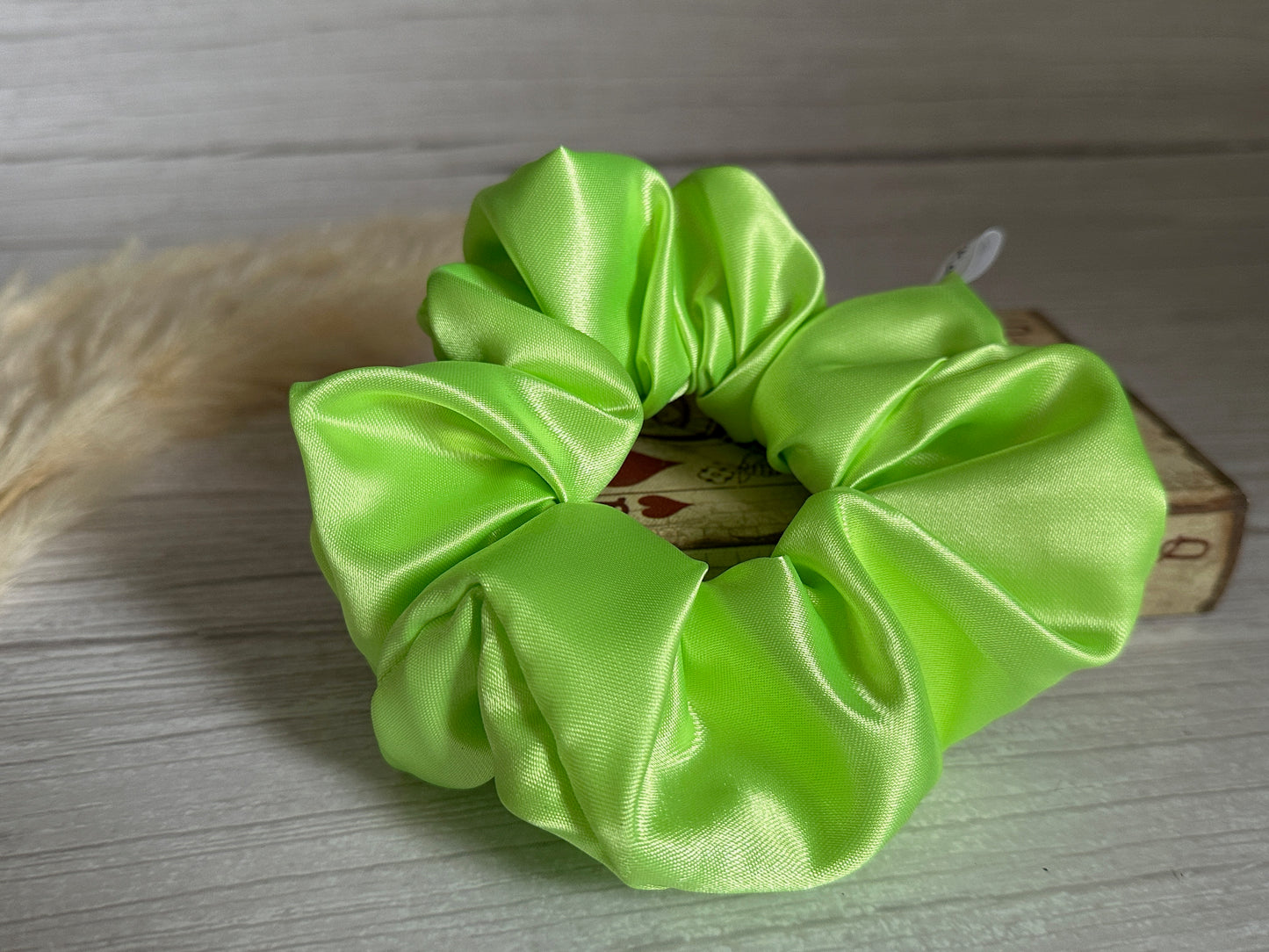 A luxurious lime green satin scrunchie from Crafts by Kate, known as the Silk Extra Large Scrunchie, graces a wooden surface. Just behind, a small box adorned with playing card designs peeks out, while to its left, a soft beige feather adds an elegant touch.