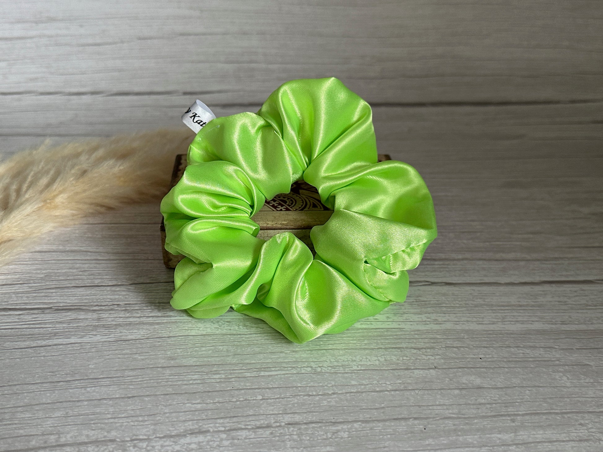 A luxurious lime green Silk Extra Large Scrunchie from Crafts by Kate rests elegantly on a gray wooden surface, complete with a small tag. A fluffy, light-colored decorative element is partially visible on the left, adding a touch of grace to this exquisite accessory.