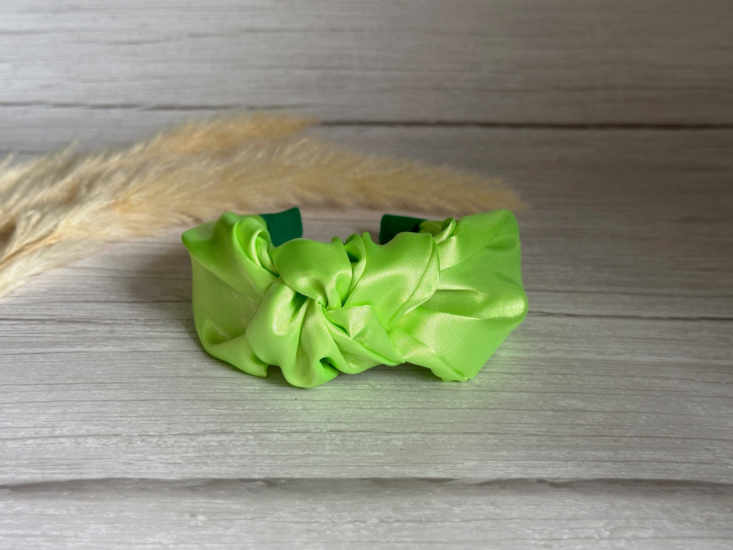 A luxurious lime green Silk Knotted Headband by Crafts by Kate rests on a light wooden surface, complemented by beige pampas grass in the corner.