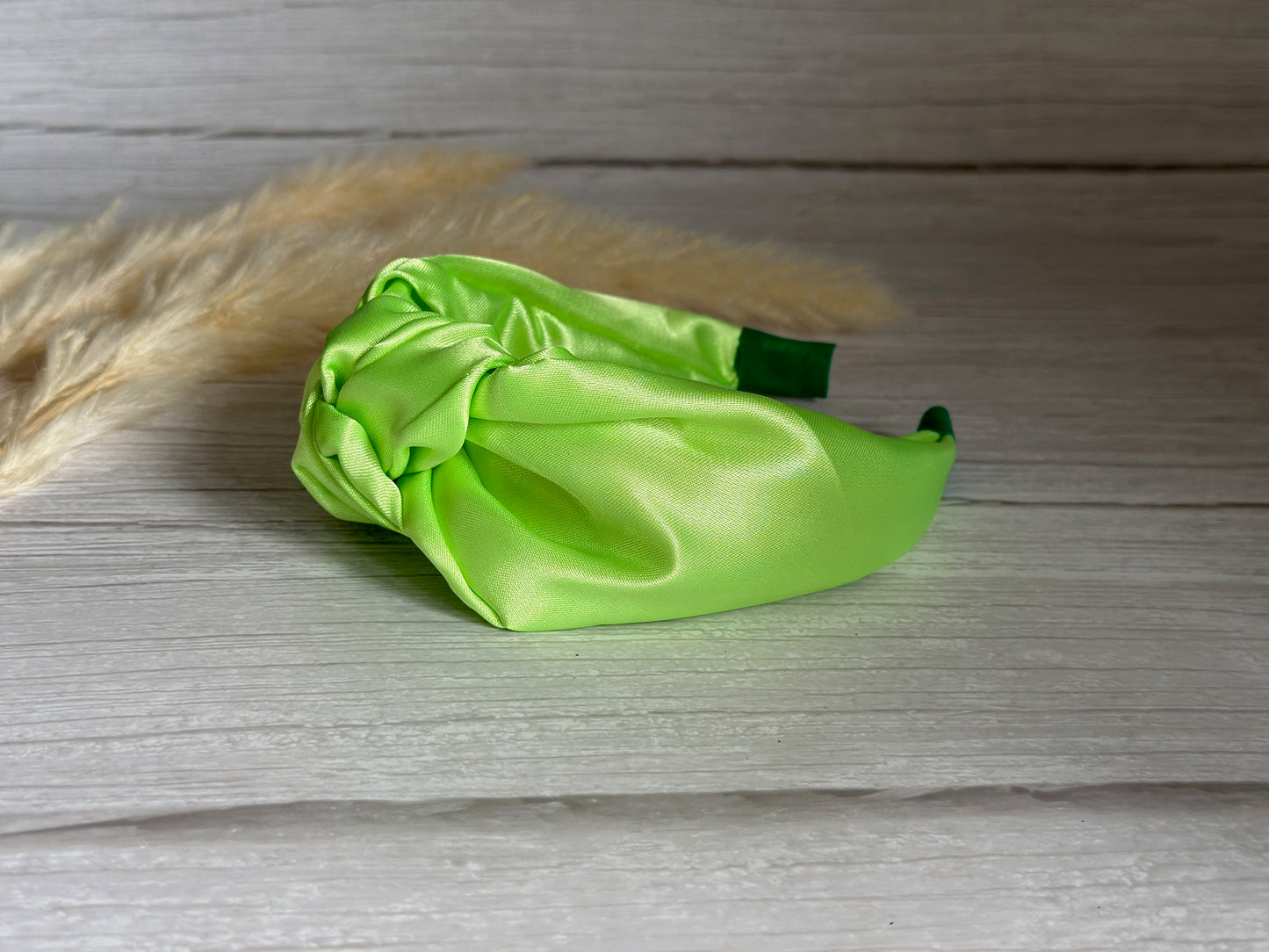 A vibrant lime green, Silk Knotted Headband by Crafts by Kate rests on a light wooden surface, with pampas grass elegantly adorning the background to add a natural touch of luxury.