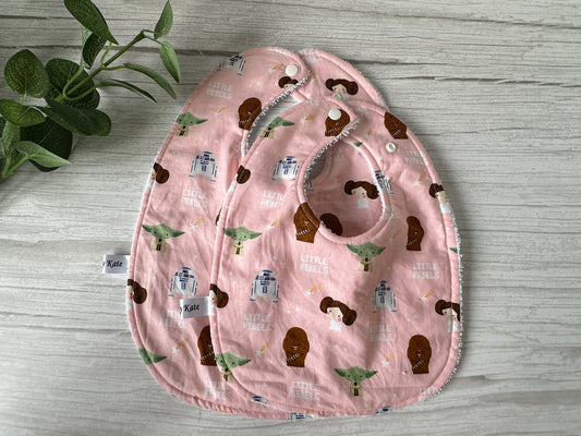 Two ultra-soft cotton bibs by Crafts by Kate, featuring the "Star Wars Little Rebels" theme, showcase delightful sci-fi character illustrations such as a green creature, robots, and a princess with buns. A handcrafted charm is intricately detailed alongside a green plant decorating the top left corner on a light wooden surface.