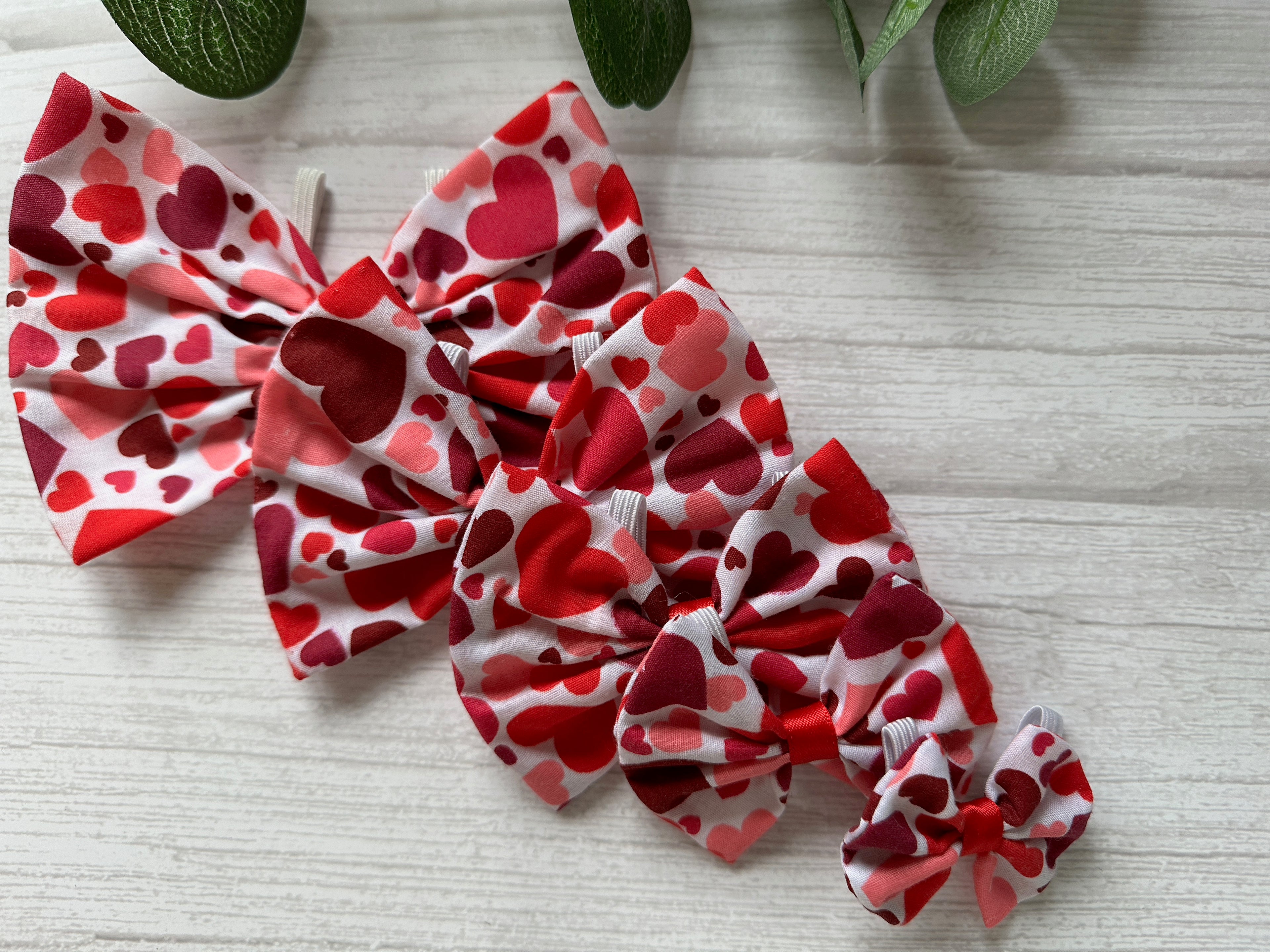 Four heart-patterned bows of different sizes, made from Crafts by Kate's Valentines Hearts cotton fabric, are arranged on a light wood surface. With elastic fastenings, these charming pet accessories with collar attachments are perfect for any occasion. Green leaves top the image.