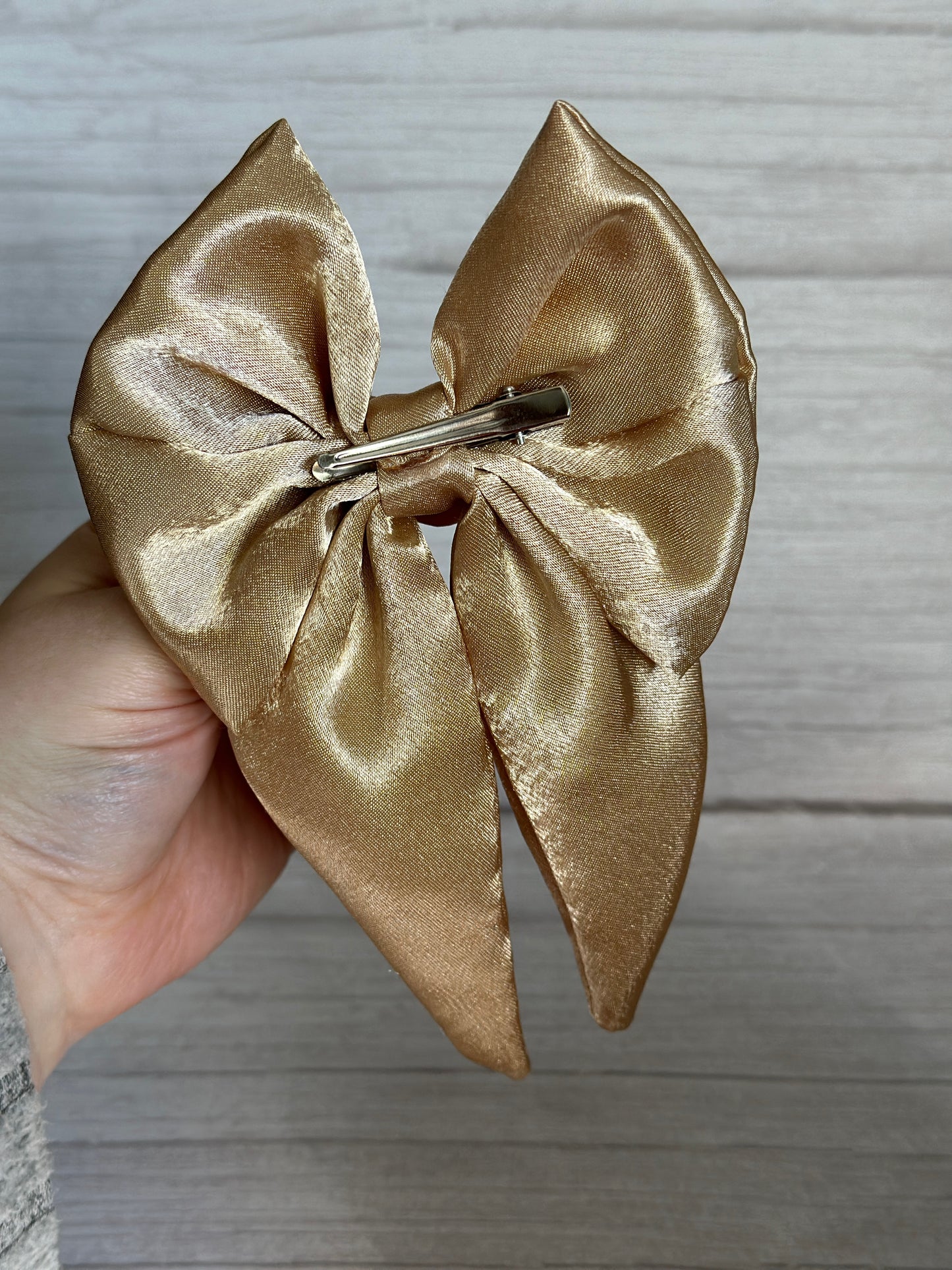 A hand is holding a large, shiny Silk Sailor Bow Clip - Luxury Gold by Crafts by Kate, featuring a handcrafted design with a metal clip on the back, set against a light wooden background.