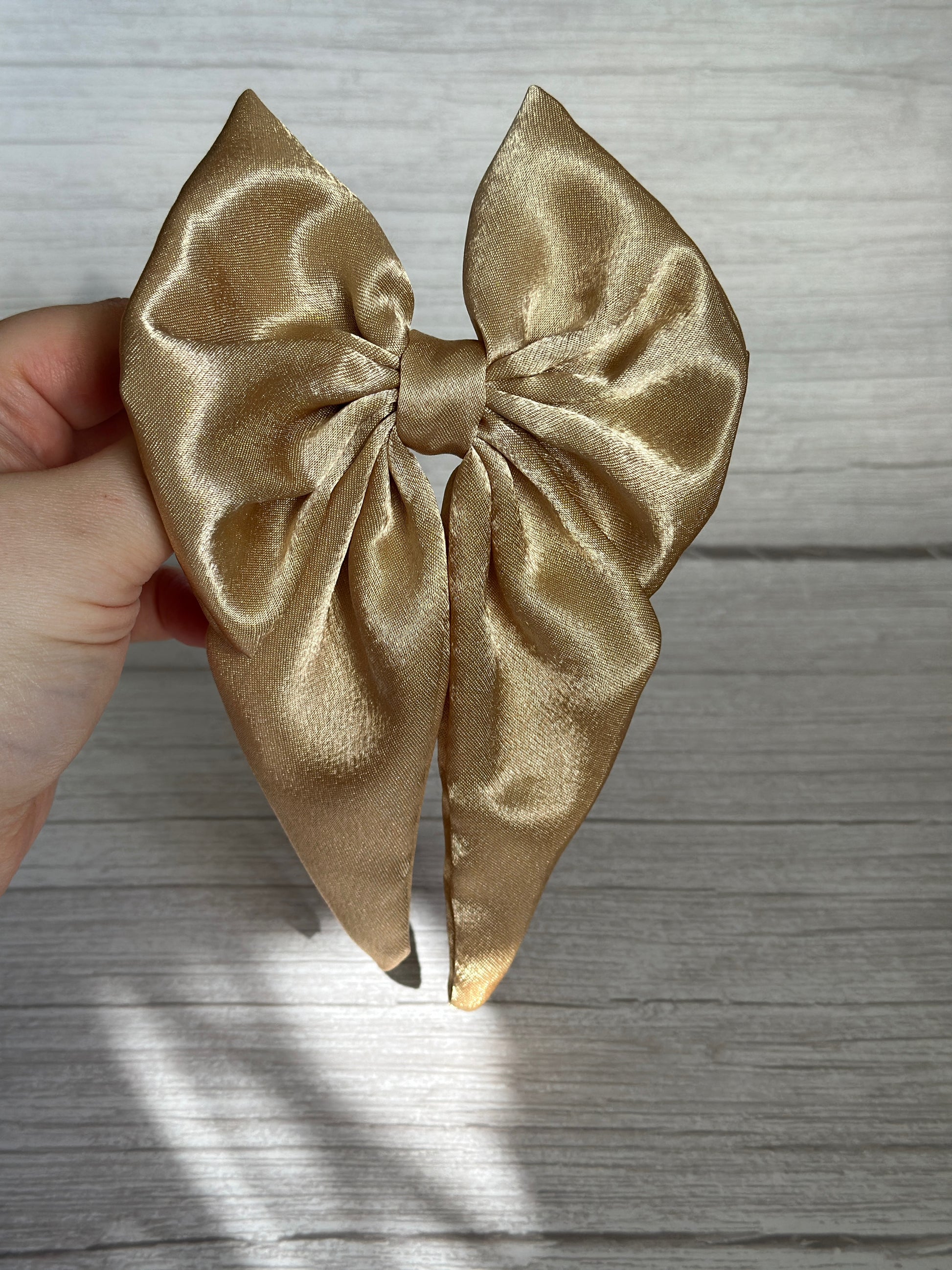 A hand holds the Silk Sailor Bow Clip - Luxury Gold from Crafts by Kate against a light gray wooden background. This handcrafted statement piece showcases two elegant loops and long trailing ribbons. Light gently casts shadows across its surface, enhancing its luxurious appeal.