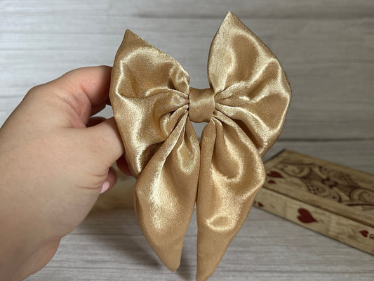 A hand presenting the luxurious Silk Sailor Bow Clip in gold from Crafts by Kate against a light wooden backdrop, with a decorative heart-themed wooden box visible in the background.