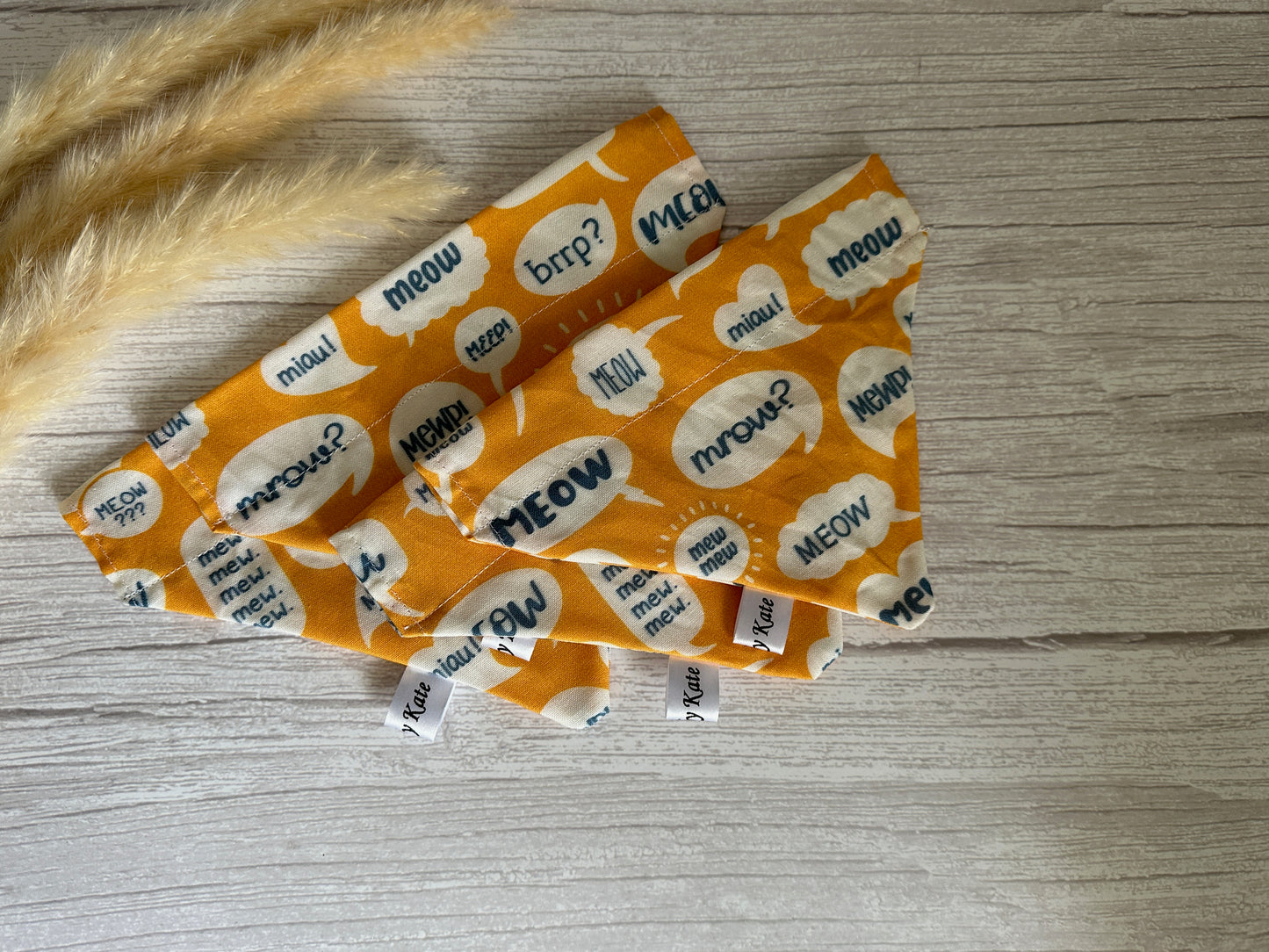 Crafts by Kate's Cotton Pet Bandana with collar attachment features Meow Fabric in orange and white cat-themed squares with "meow" speech bubbles, set on wood next to three pampas grass pieces, ideal for crafting.