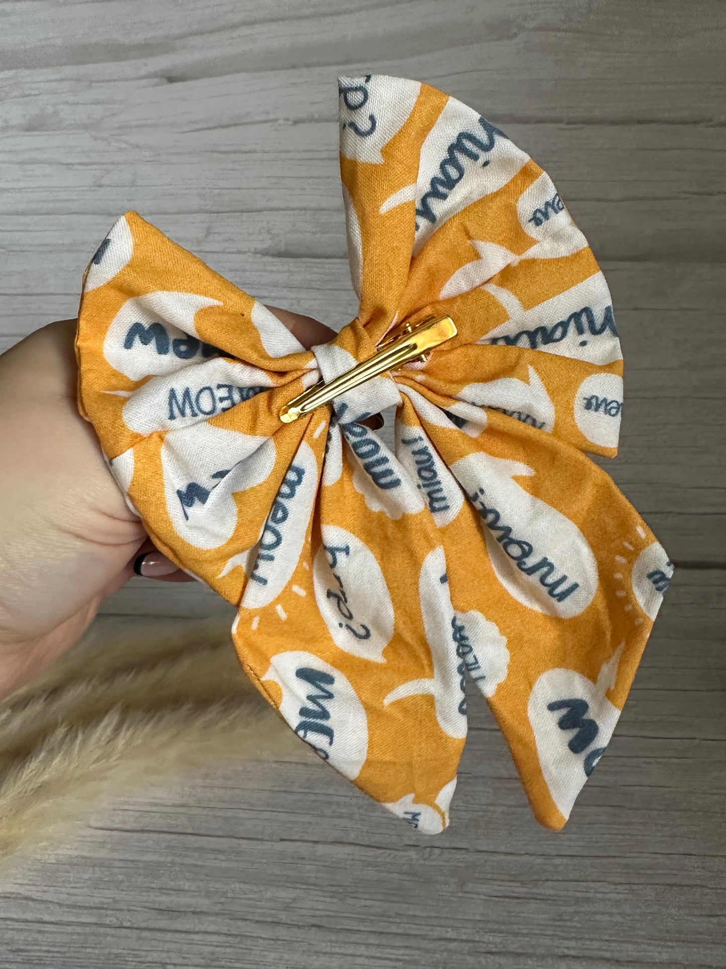 A person holds the "Cotton Sailor Bow Clip - Meow" by Crafts by Kate, a striking luxury statement piece in vibrant orange fabric, featuring playful speech bubbles with "meow" and "me?" against a wooden background. The bow is elegantly secured with a gold crocodile clip.