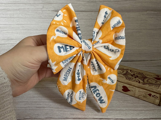 A hand holds the luxurious orange Cotton Sailor Bow Clip - Meow by Crafts by Kate, adorned with white speech bubbles saying "MEOW" in blue text. This bow, attached with a crocodile clip, is displayed against a wooden surface alongside a decorative box embellished with hearts and ornate designs.