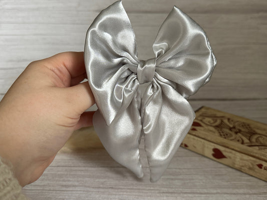 A hand displays a beautifully crafted Silk Sailor Bow Clip in metallic silver from Crafts by Kate against a light wooden background. The bow features long tails and a smooth, shiny texture. A wooden box adorned with heart designs is partially visible, adding to the handcrafted luxury appeal.