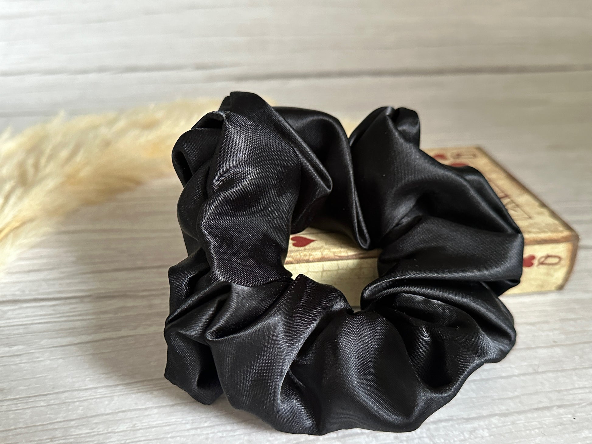 A Silk Extra Large Scrunchie - Midnight Black by Crafts by Kate rests on a small, decorative box with rustic tones. The scrunchie's smooth texture contrasts dramatically with the box's vintage look, set against a light wooden background. A soft, beige pampas grass plume adds to the handcrafted luxury vibe beside it.