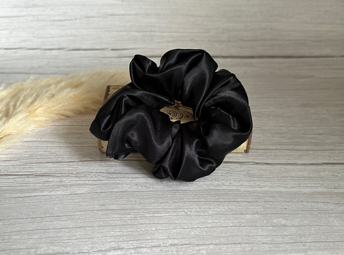 A Silk Extra Large Scrunchie in Midnight Black from Crafts by Kate is elegantly displayed on a small wooden block against a light gray wood background. To the left, a piece of cream-colored, dried foliage peeks into view, enhancing the luxurious feel.