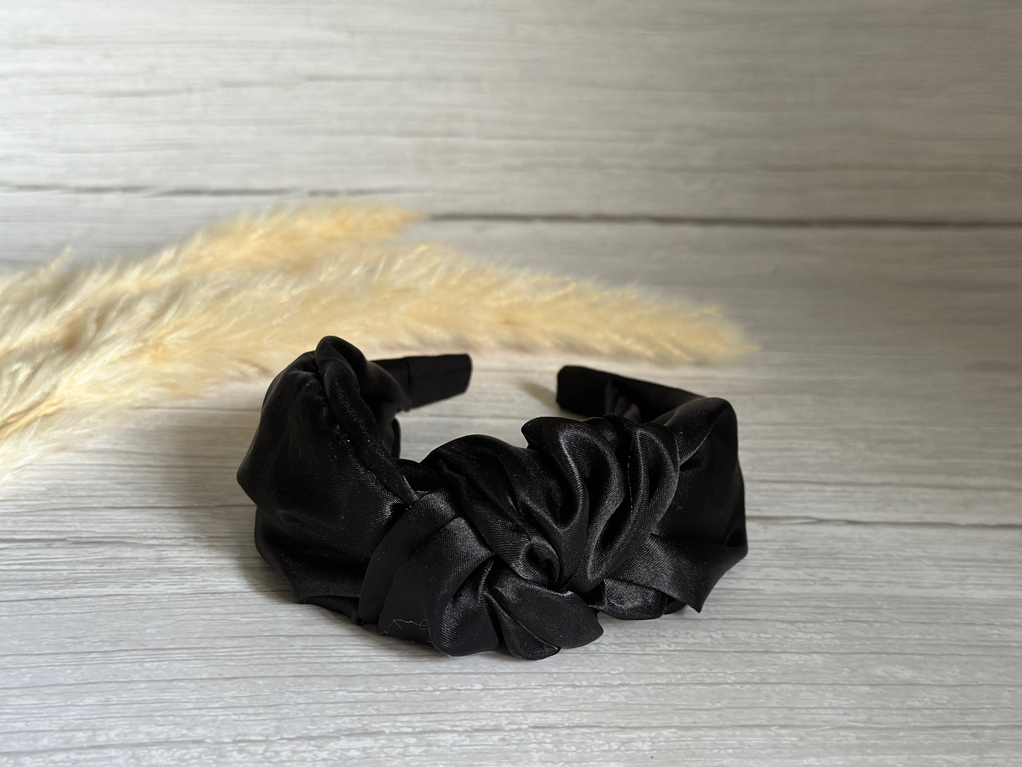 A Silk Knotted Headband in Midnight Black from Crafts by Kate rests on a light wooden surface, its ruched design standing out. In the background, soft beige pampas grass adds a delicate and natural contrast, enhancing the luxurious ambiance.