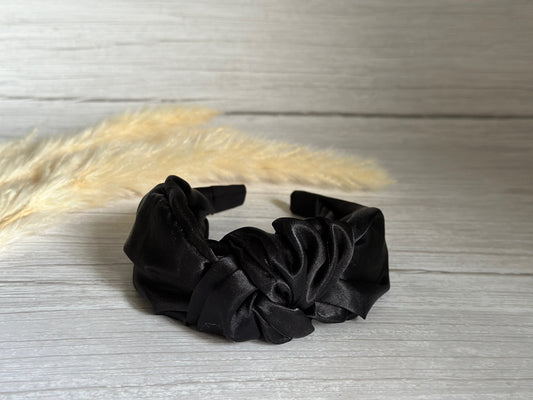 A Silk Knotted Headband in Midnight Black from Crafts by Kate rests on a light wooden surface, its ruched design standing out. In the background, soft beige pampas grass adds a delicate and natural contrast, enhancing the luxurious ambiance.