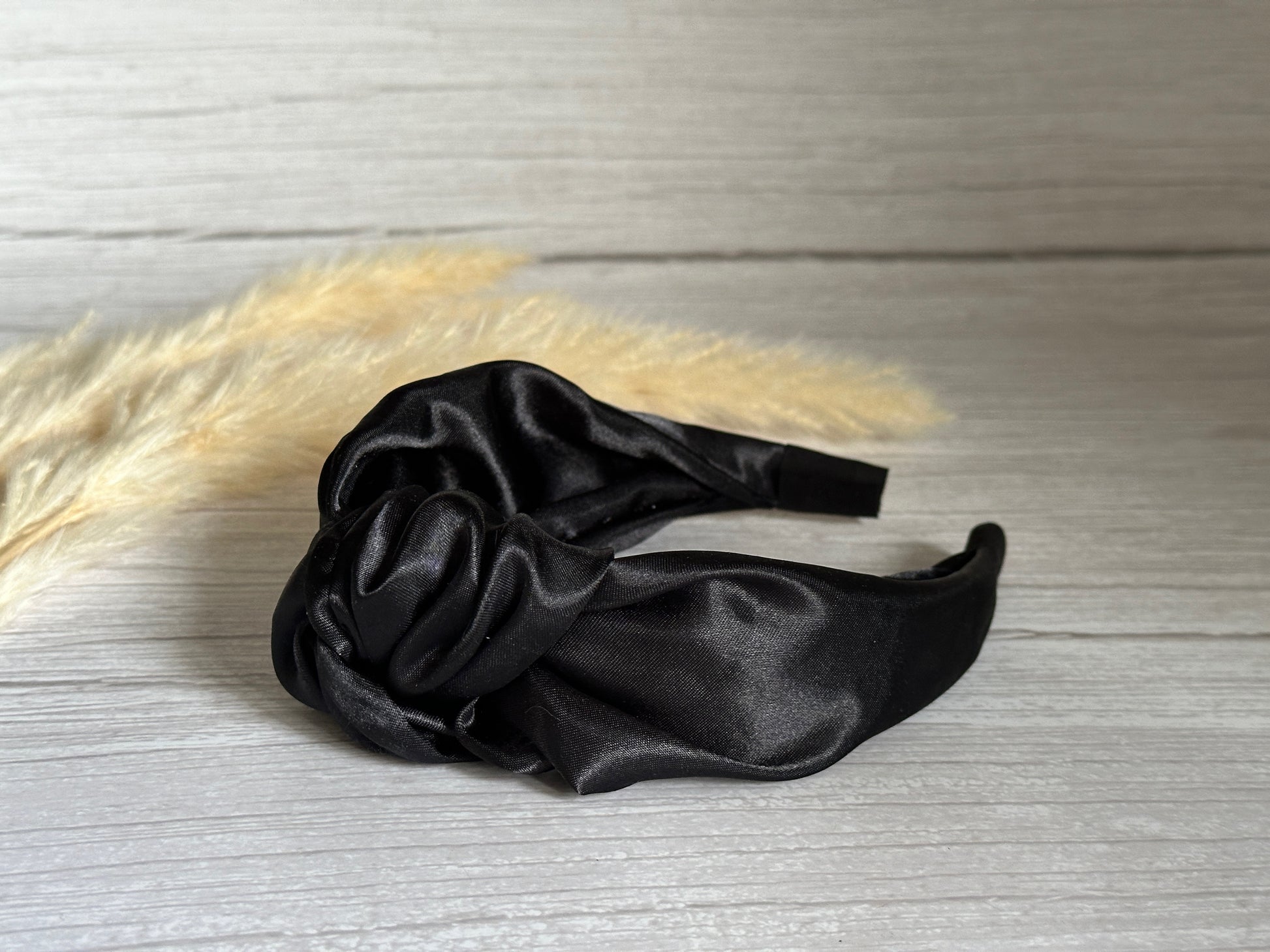 The Silk Knotted Headband - Midnight Black by Crafts by Kate rests elegantly on a light wooden surface, its luxurious satin finish and knotted design radiating sophistication. In the softly blurred background, beige dried grass stems add a touch of natural texture.