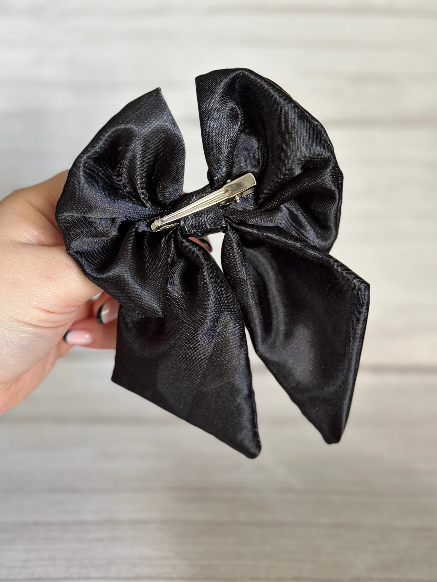 A hand holds the Crafts by Kate Silk Sailor Bow Clip in Midnight Black, featuring a handcrafted statement bow with a metal barrette on the back. The backdrop is a light, textured surface, accentuating the elegant design of the luxury silk bow.