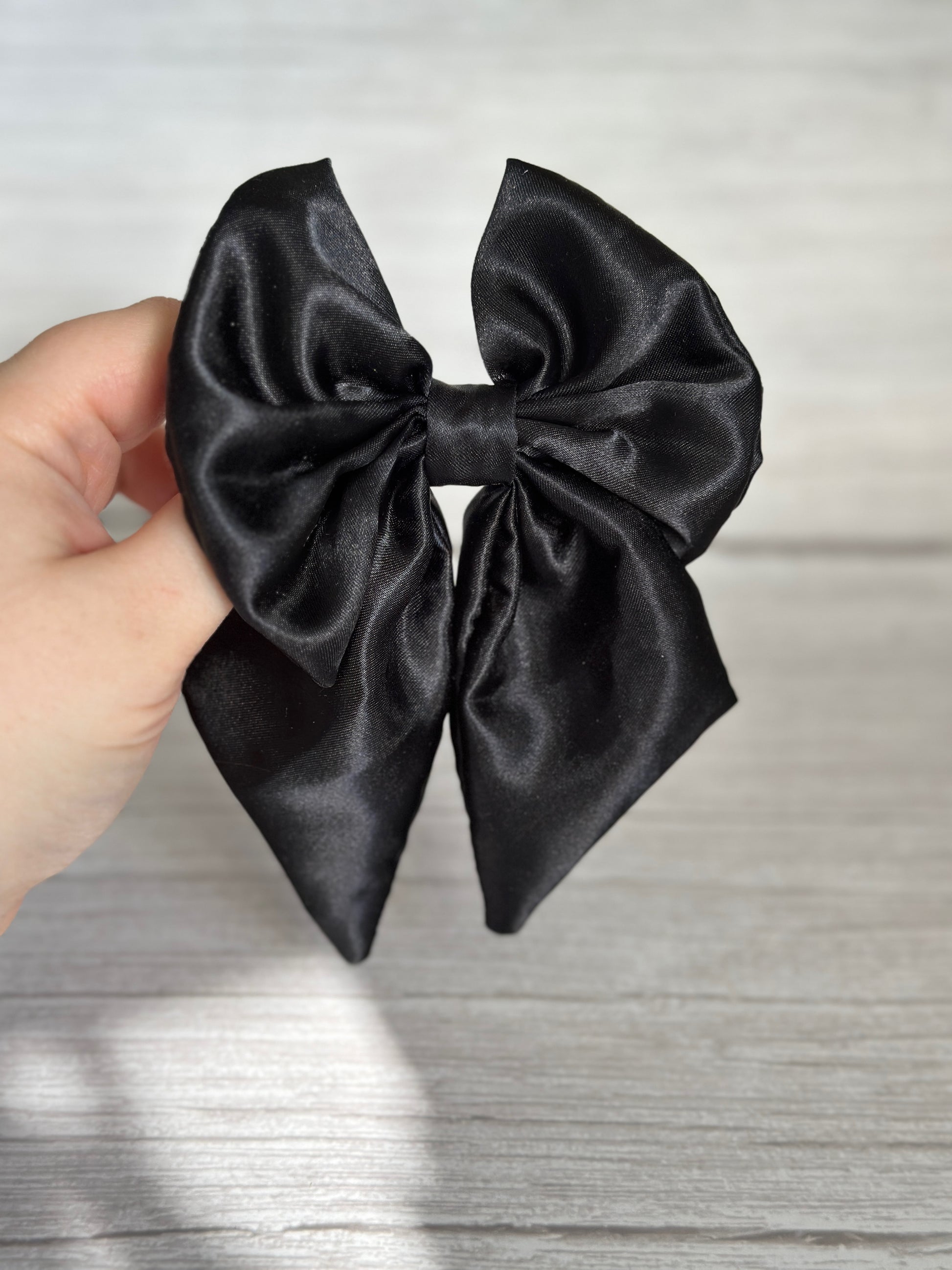 A hand holding a luxurious Midnight Black Silk Bow Clip from Crafts by Kate against a light wooden background, showcasing its elegance and timeless charm.