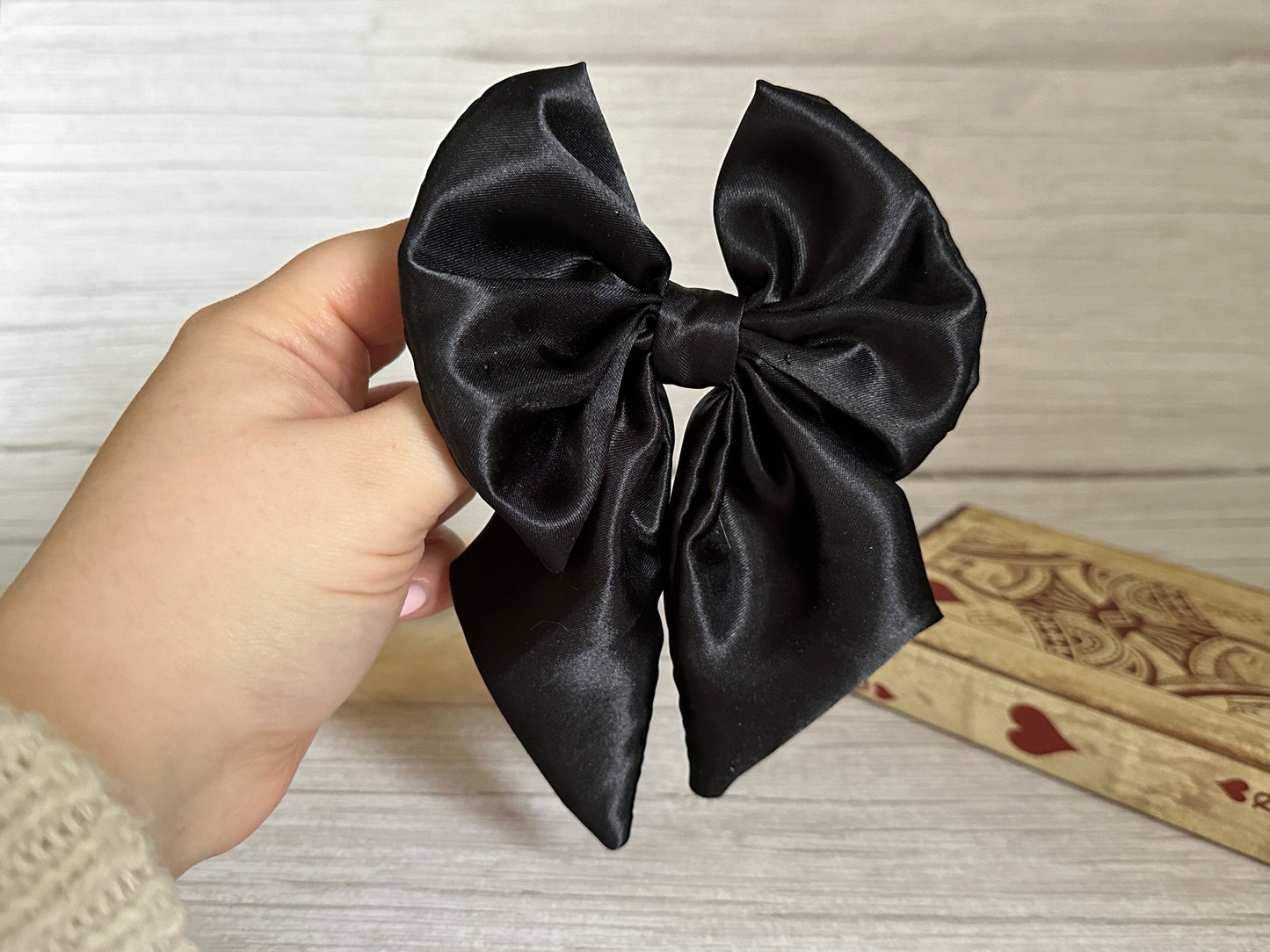 A hand is holding the Crafts by Kate Silk Sailor Bow Clip in midnight black, showcasing its elegant craftsmanship against a light wooden background. On the right, a decorative wooden box adorned with heart patterns is partially visible.