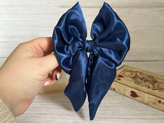 A hand holding a luxurious Silk Sailor Bow Clip in Midnight Blue against a light wood background. A wooden box adorned with heart designs from Crafts by Kate is partially visible on the side.