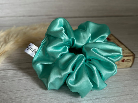 A luxurious mint green Silk Extra Large Scrunchie by Crafts by Kate is elegantly displayed on a light wooden surface. It features a small white tag, and the background showcases soft fur alongside a decorative box adorned with hearts.