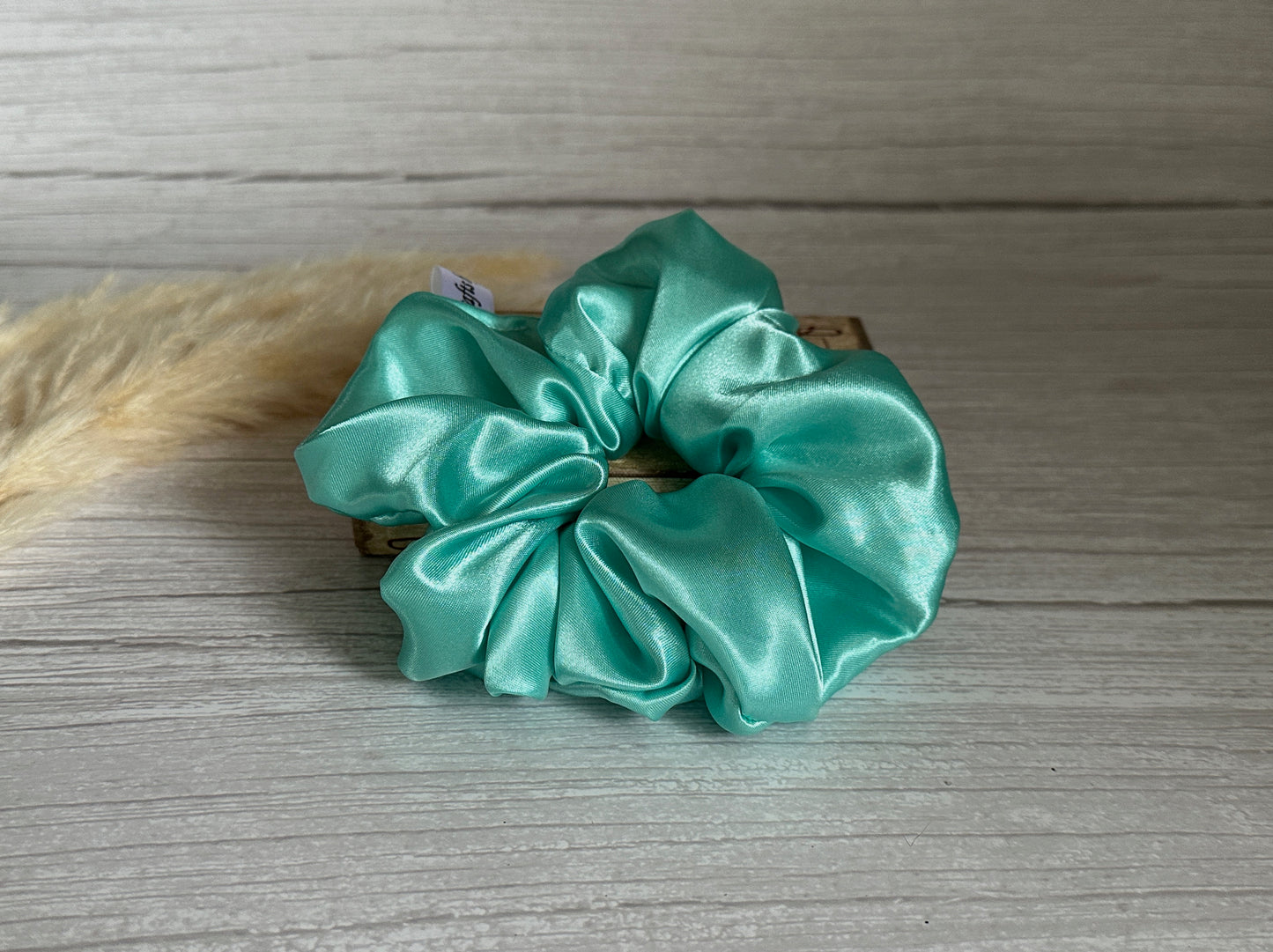 A luxurious Silk Extra Large Scrunchie in mint green by Crafts by Kate graces a light wooden surface, with a fluffy beige object partially visible in the background.