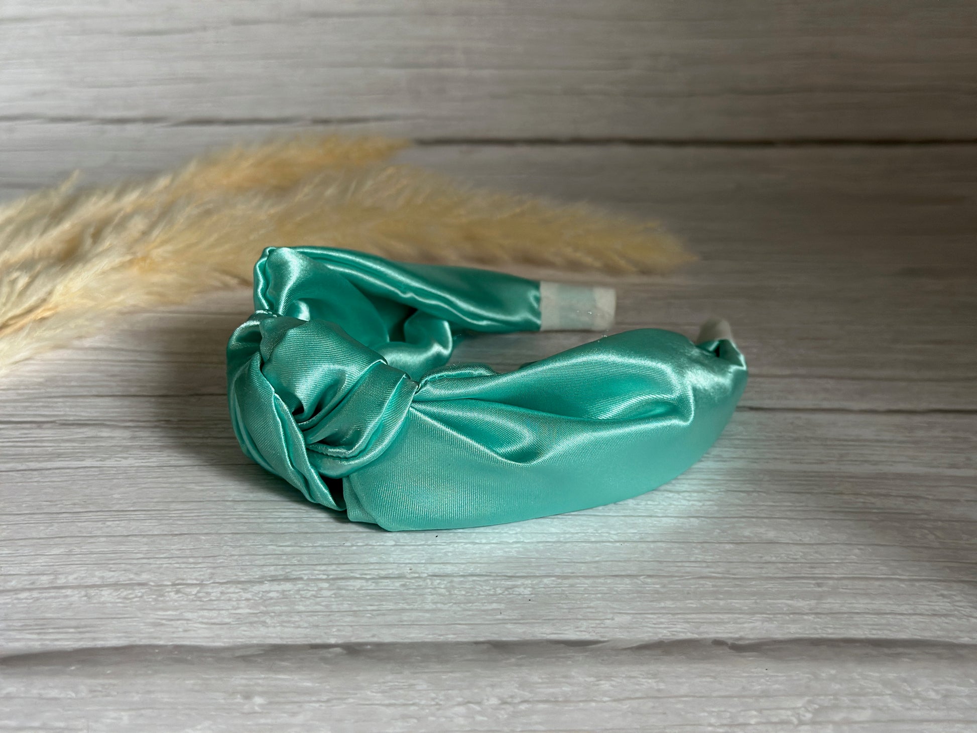 The Silk Knotted Headband - Mint Green by Crafts by Kate features a luxurious twisted knot design, elegantly displayed on a light wooden surface. Softly blurred pampas grass in the background enhances its sophisticated allure.