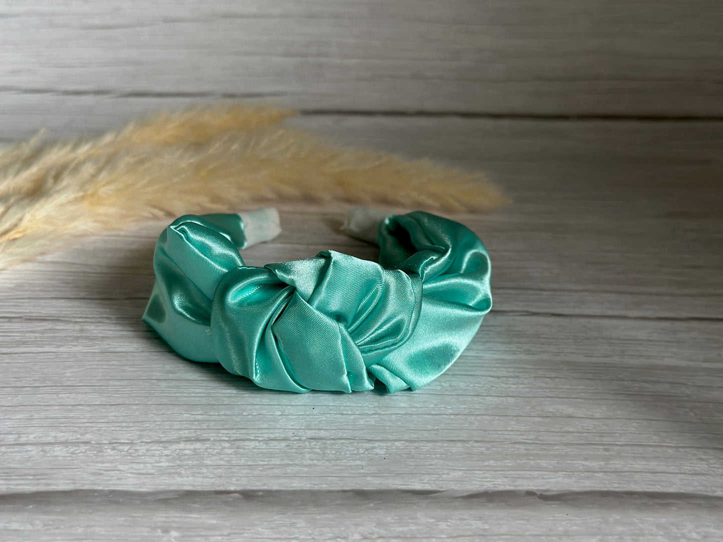 The Silk Knotted Headband - Mint Green by Crafts by Kate lies gracefully on a light wooden surface, with soft beige pampas grass creating a blurred background.