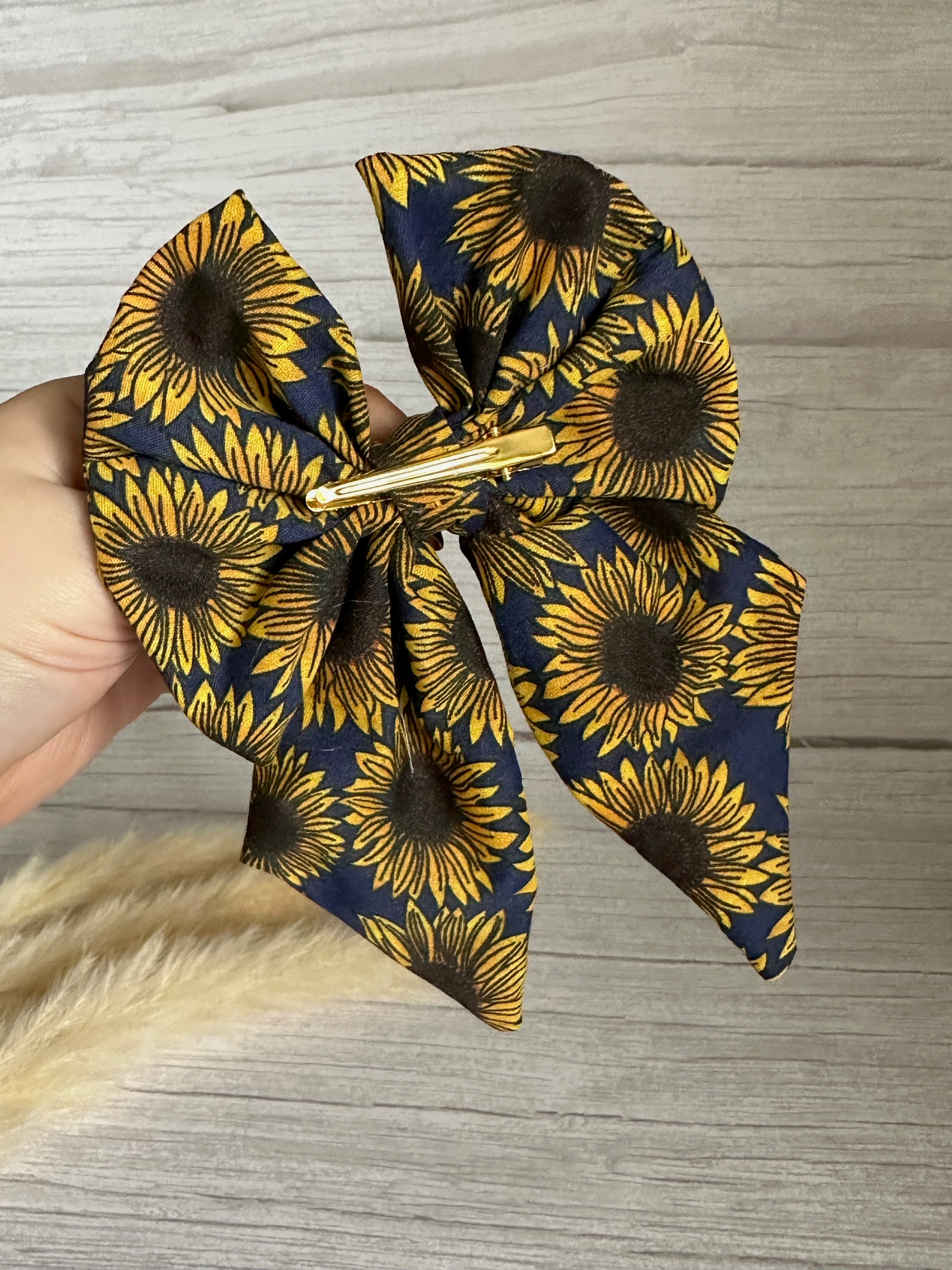 A hand holds the Cotton Sailor Bow Clip - Navy Sunflowers, a handcrafted luxury accessory by Crafts by Kate. This cotton bow features vibrant yellow sunflowers with black centers on a deep navy background. The textured light wood surface in the background enhances the bow's elegance.