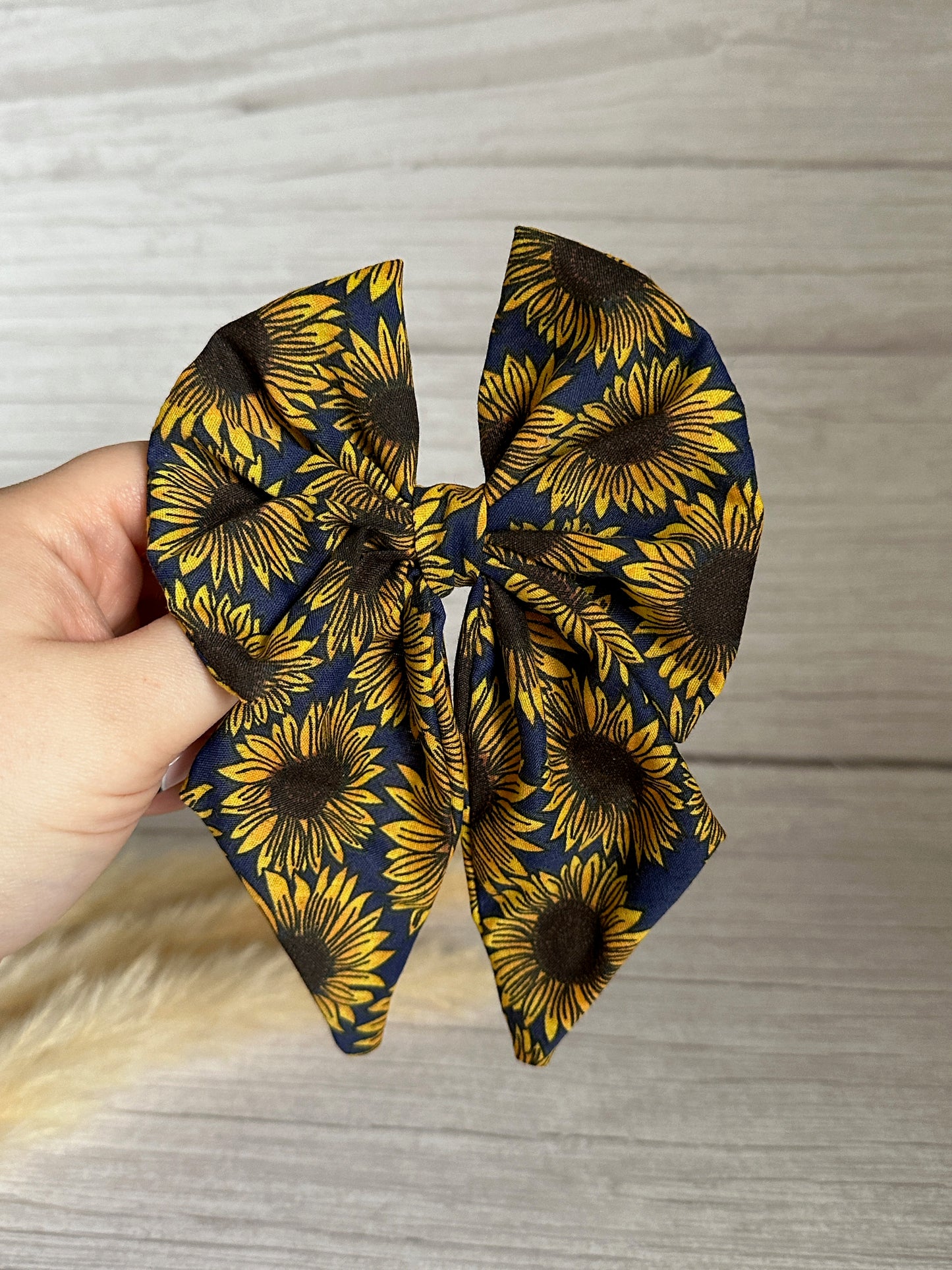 A hand holds the Crafts by Kate Cotton Sailor Bow Clip - Navy Sunflowers, crafted from luxurious cotton fabric, showcasing a vibrant sunflower pattern in yellow and brown against a dark background. The elegant bow rests gracefully on a light wooden surface.