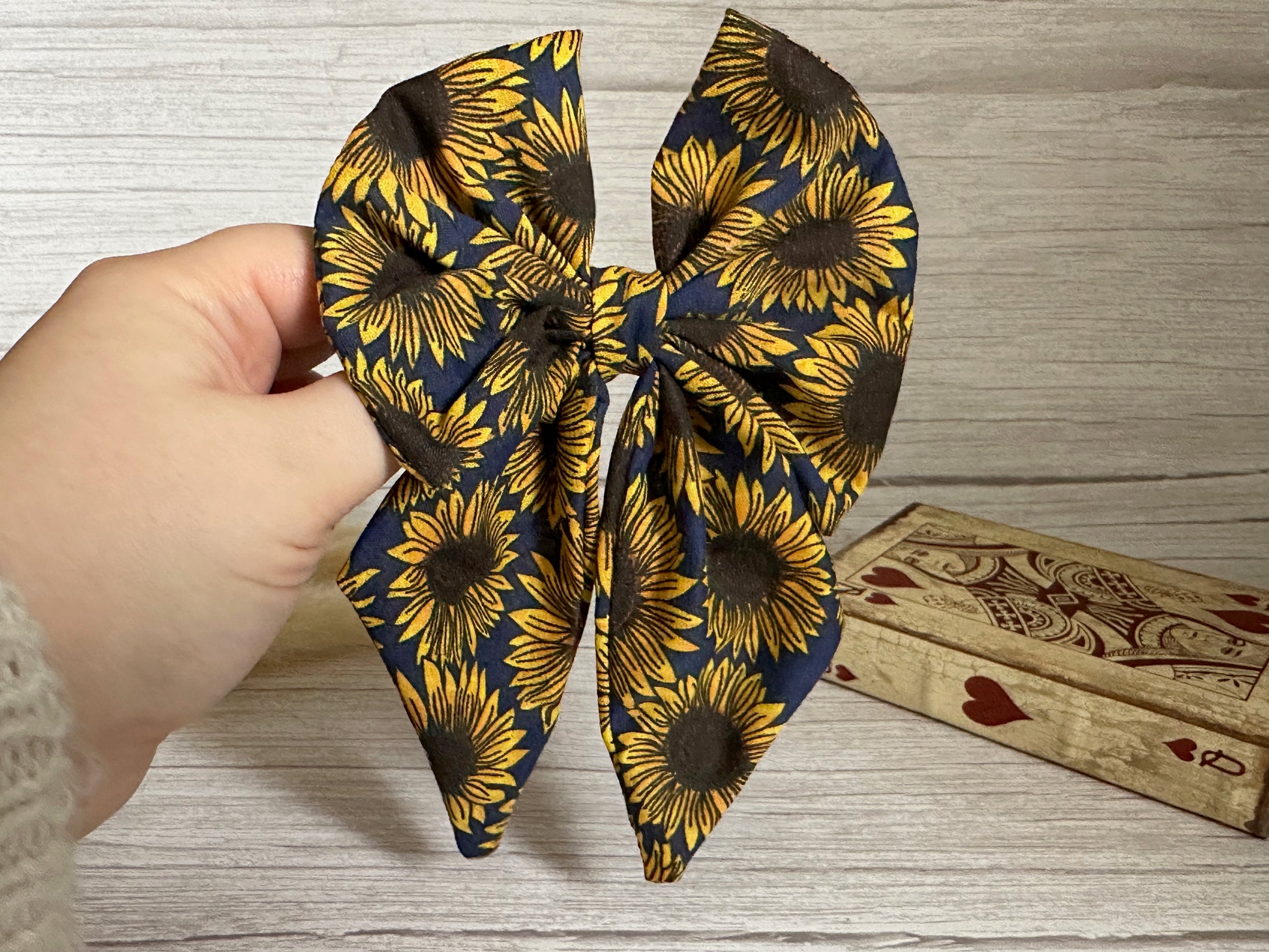 A hand displays the luxurious Cotton Sailor Bow Clip - Navy Sunflowers by Crafts by Kate, featuring a charming sunflower pattern against a wooden background. Next to it, a beautifully decorated wooden box with heart accents subtly enhances the allure of this stylish navy sailor bow.