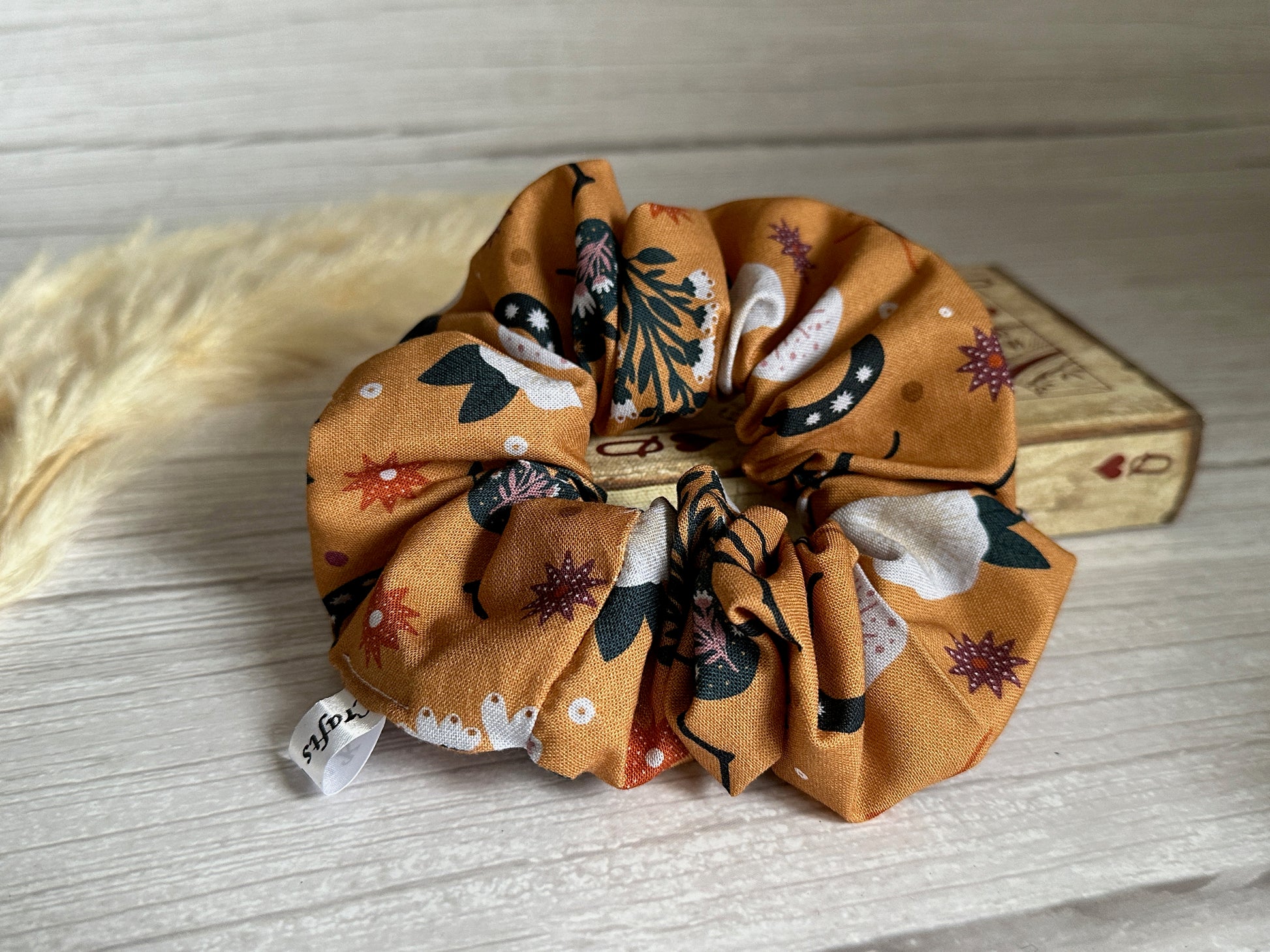 A Crafts by Kate Cotton Extra Large Scrunchie - Orange Floral, featuring a bold abstract pattern in brown, green, white, and orange, rests on a light wooden surface. Nearby lies a dried grass stem and a vintage-style wooden box, enhancing the elegant presentation of this luxury scrunchie.