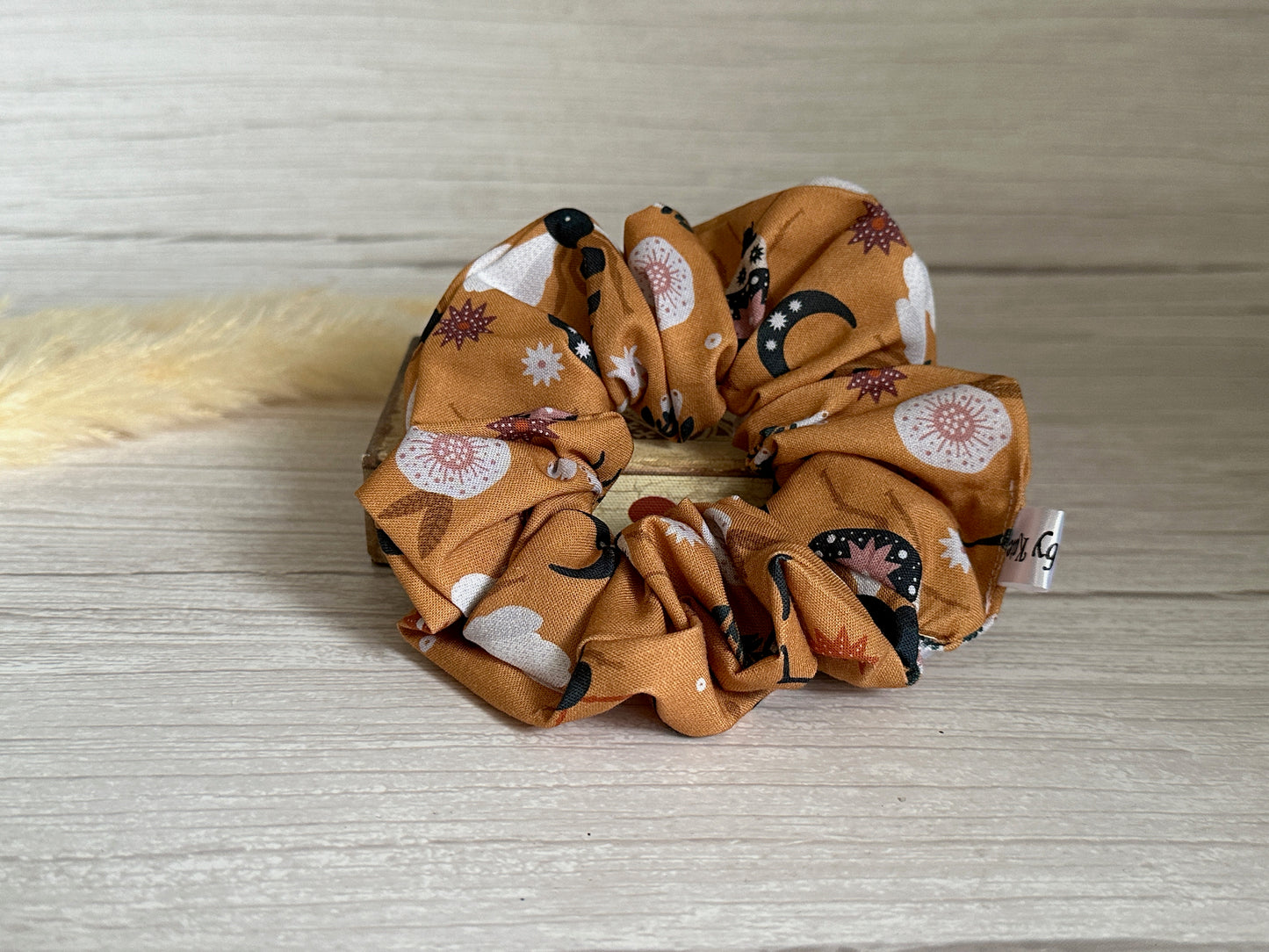 The Cotton Extra Large Scrunchie - Orange Floral by Crafts by Kate, featuring a luxurious design with an orange background and floral patterns, is elegantly placed on a light wooden surface.