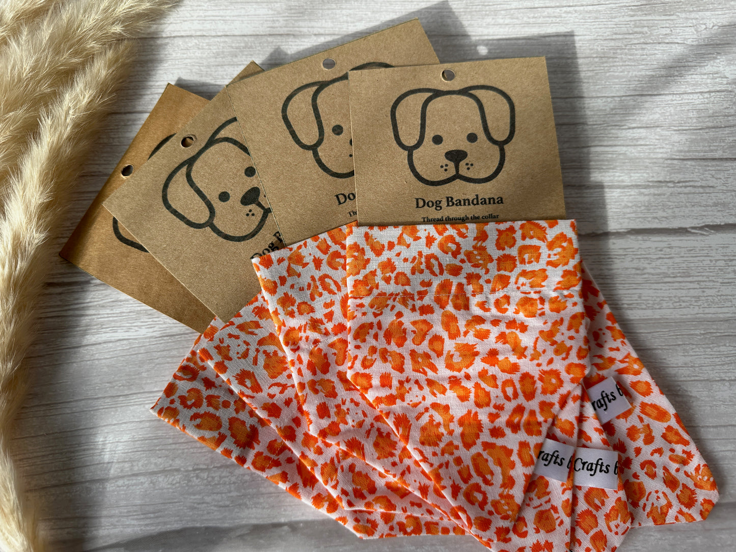 Five Cotton Pet Bandanas with collar attachments in orange leopard print, handcrafted by Crafts by Kate from soft cotton, packaged with a charming dog illustration labeled "Dog Bandana," displayed on a light wooden surface.