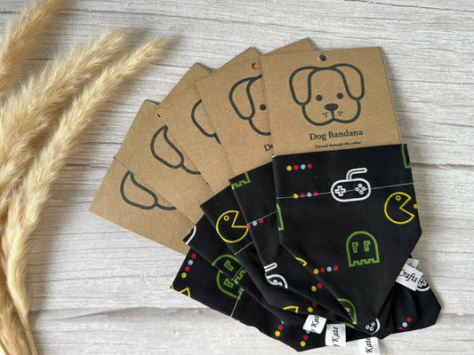 A set of five Cotton Pet Bandanas with collar attachments from Crafts by Kate, featuring retro gaming designs including a Pacman print fabric, are folded and arranged in a fan pattern. Each bandana comes with a kraft paper tag adorned with a simple dog illustration and the text "Dog Bandana." The bandanas and tags rest on a light wood surface next to dried wheat.