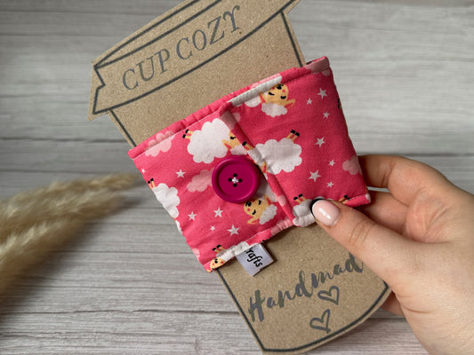 A person holds a Cotton Reusable Cup Cozy - Pink Sheep by Crafts by Kate. This eco-friendly accessory, made from pink fabric adorned with cute lambs, clouds, and stars, features a pink button for securing it around a cup. It is attached to cardboard backing labeled "Cup Cozy" with "Handmade" written at the bottom.